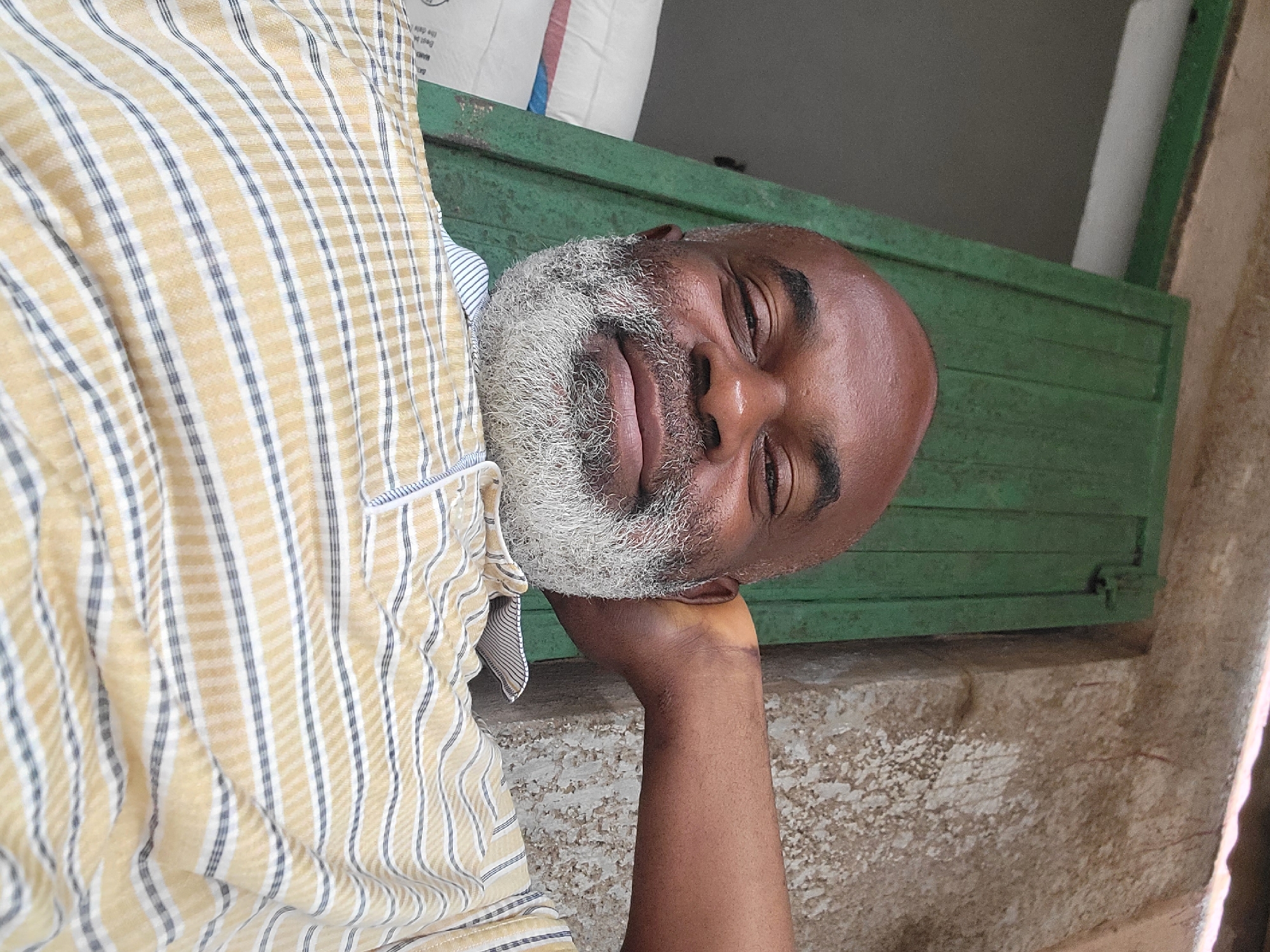 An adult Black male with a bald head and a grey beard is reclining with his head resting on his right hand. He is wearing a yellow and white striped shirt. Behind him is a green painted wooden structure, possibly a window or door frame, and a textured surface that appears to be a wall or ledge. A bag of flour is leaning against the structure. He is located in the city of Maiburiji, Nigeria.