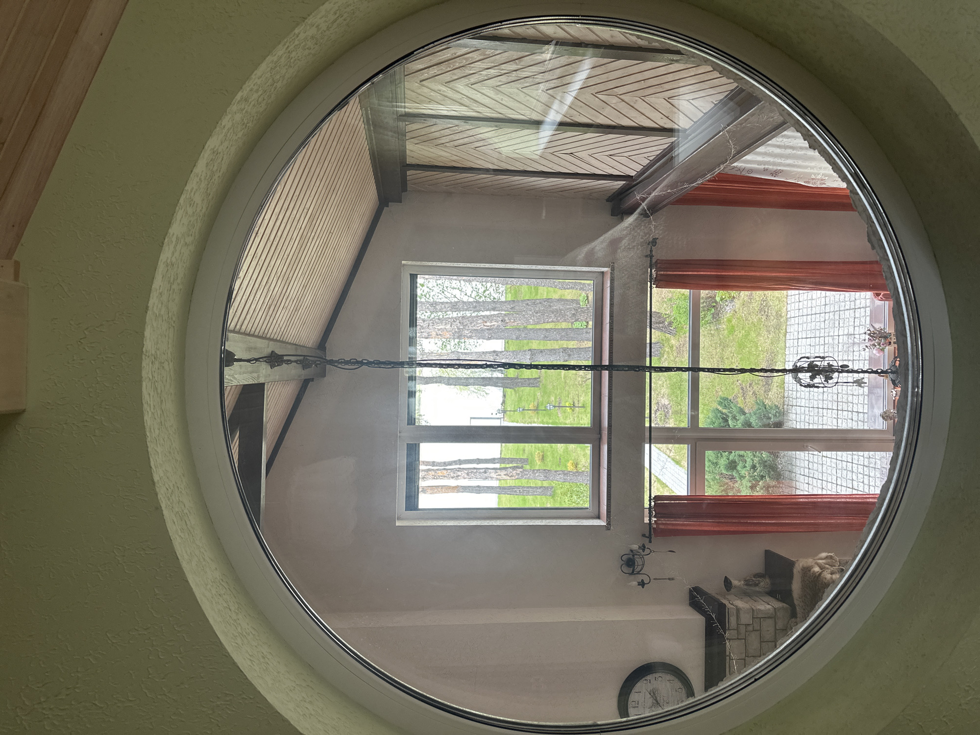 The image shows an interior view through a circular window. Reflected in the window's glass is the interior of a room. A rectangular window is visible behind the circular one, showing trees and a grassy landscape. In the reflected room, there's a fireplace with a cat on top, curtains, and a clock on the wall. A chandelier hangs from the ceiling, also reflected. The exterior frame of the circular window appears to be white and is mounted on a textured wall, which is yellow. The presence of certain materials and items, such as the clock and fireplace, suggests this could be a residential setting. The location is Aviator, Russia.