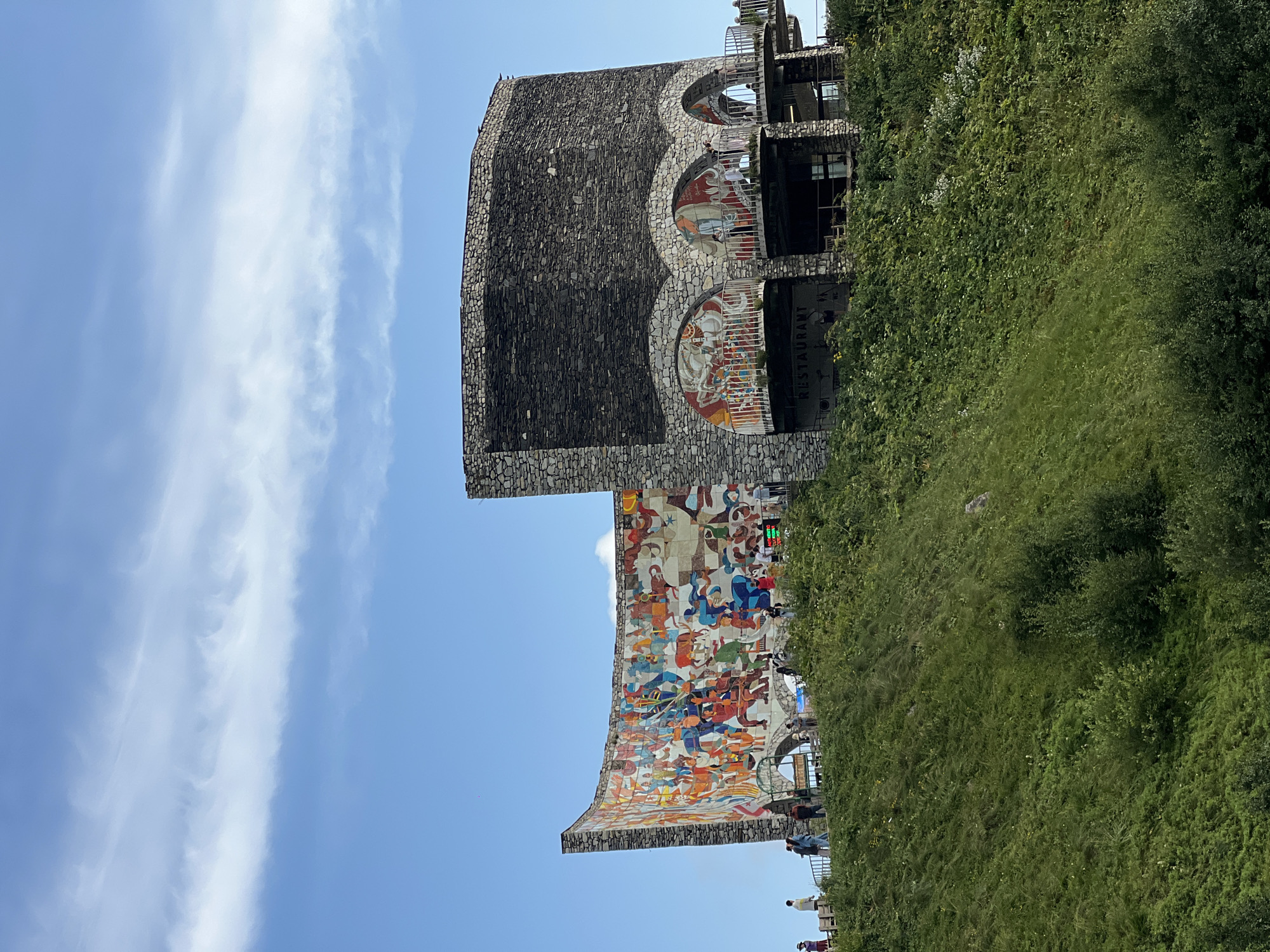 The image captures the Gudauri Friendship Monument, a striking circular-shaped structure built from stone, situated on a mountain slope in Gudauri, Georgia. The monument features a large, vibrant mural depicting historical scenes and figures, which adorns a curved wall on one side. Adjacent to this is a cylindrical tower, also constructed of stone, with arched openings that reveal balconies and what appears to be a restaurant area. Several people are visible on the walkways and the elevated platform of the monument, some standing and observing the scenery, while others are captured in motion. The monument is surrounded by lush green vegetation that covers the steep hillside, under a bright blue sky with scattered wispy clouds.
