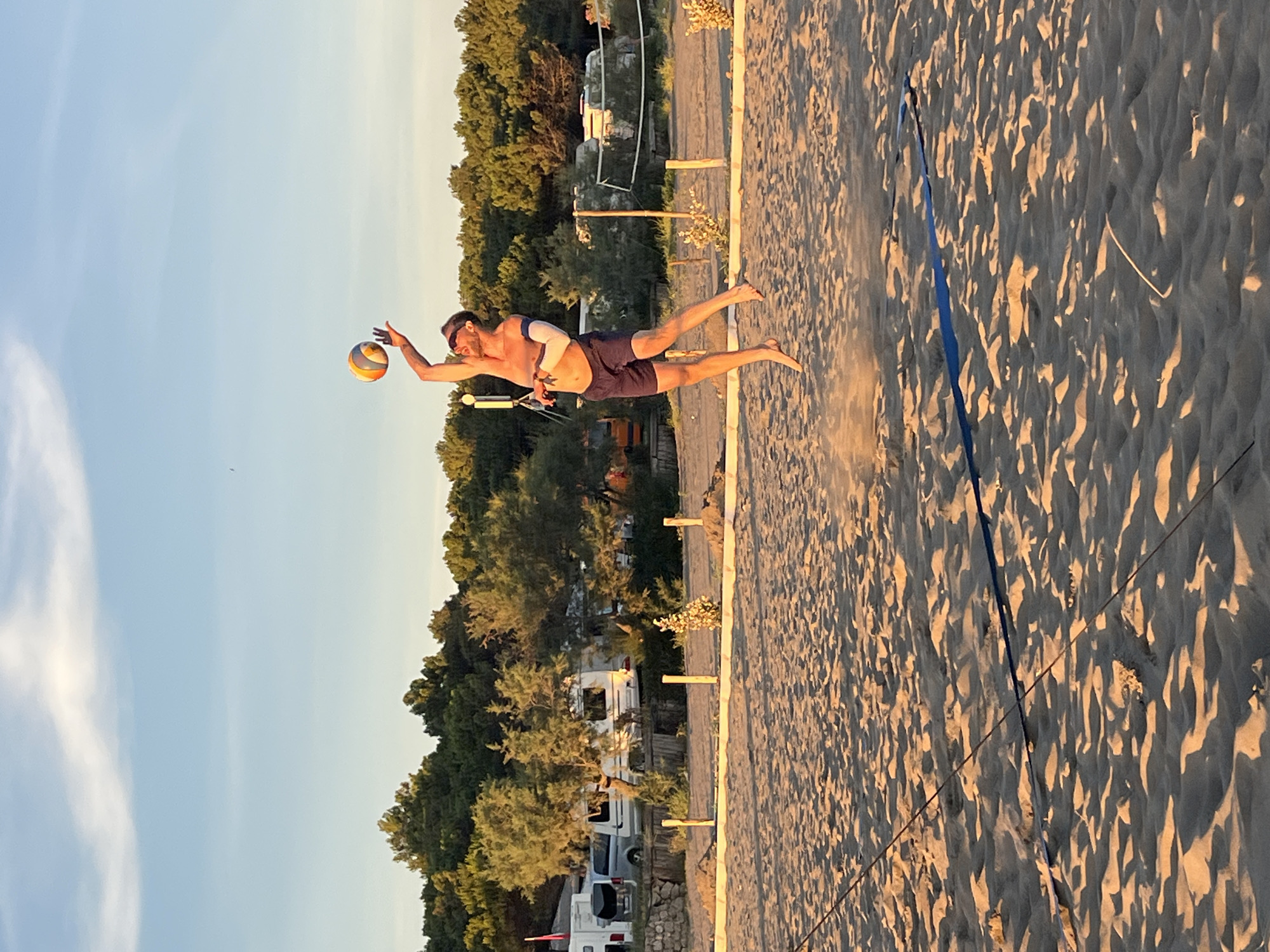 A man is playing beach volleyball at sunset in what appears to be a campground near Pistula, Montenegro. He is in mid-air, jumping to hit the ball, which is above his head and slightly to his left. He is wearing dark blue shorts and has a blue arm brace on his left arm. The sun is setting, casting long shadows across the sandy beach. In the background, there are trees, some parked RVs, and a volleyball net. The sky is clear with a few wispy clouds. The overall scene is one of recreation and leisure.
