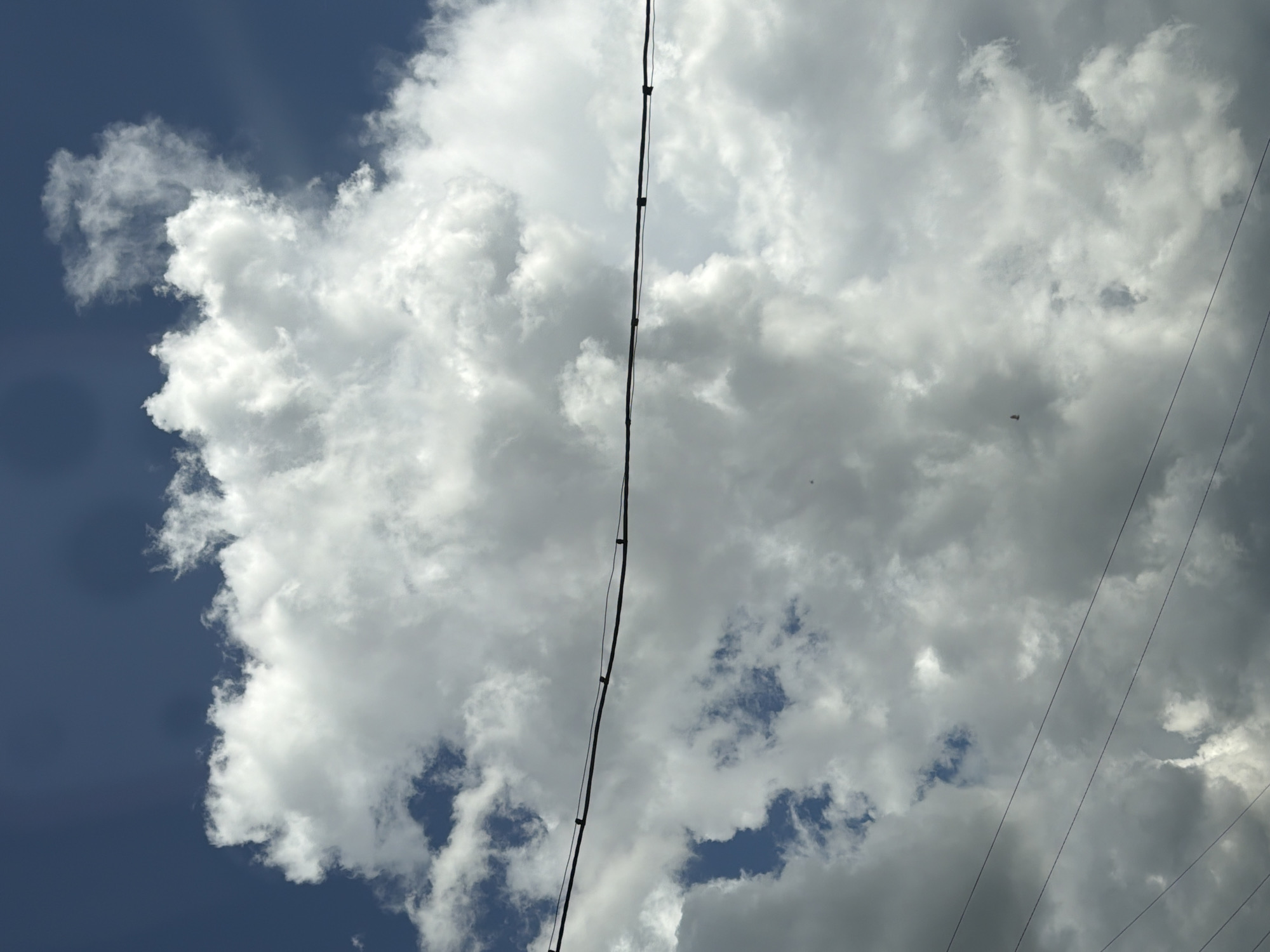 The image shows a dark blue sky with large, fluffy white clouds. A black wire stretches horizontally across the middle of the frame, supported by dark vertical posts. There are also several thinner wires running diagonally across the lower portion of the image. The overall impression is of an overcast sky with some areas of bright sunlight breaking through. The scene appears to be an outdoor setting, possibly in a rural or suburban area, given the presence of overhead wires. The image does not provide any specific clues about the city of Mezhevoy, Russia, or any other location.