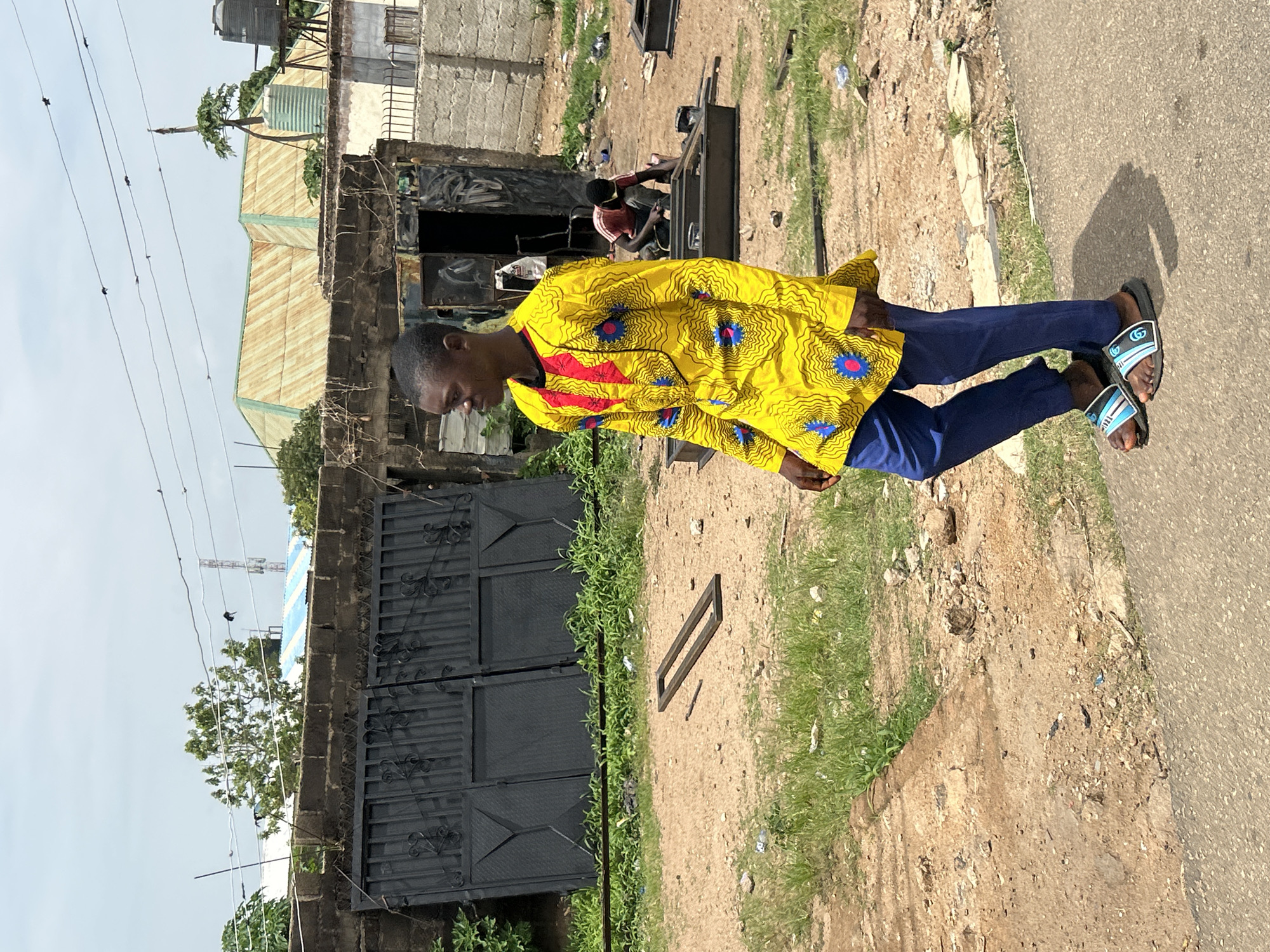 A medium shot captures a young Black man walking to the right in the foreground. He wears a bright yellow, long-sleeved shirt with a red V-neck detail and a blue and pink floral pattern. His pants are a dark blue, and he wears black and blue striped slides. The man is on a paved surface, with dry, dusty ground and patches of green grass surrounding him. In the background, a large black metal gate stands open, revealing a dark interior. Beyond the gate, a man in a red shirt and dark pants appears to be working on metal structures. Several corrugated metal structures with rounded roofs are visible further in the distance, along with trees and utility poles. The sky is overcast and bright. This scene is located in the city of Jos, Nigeria.