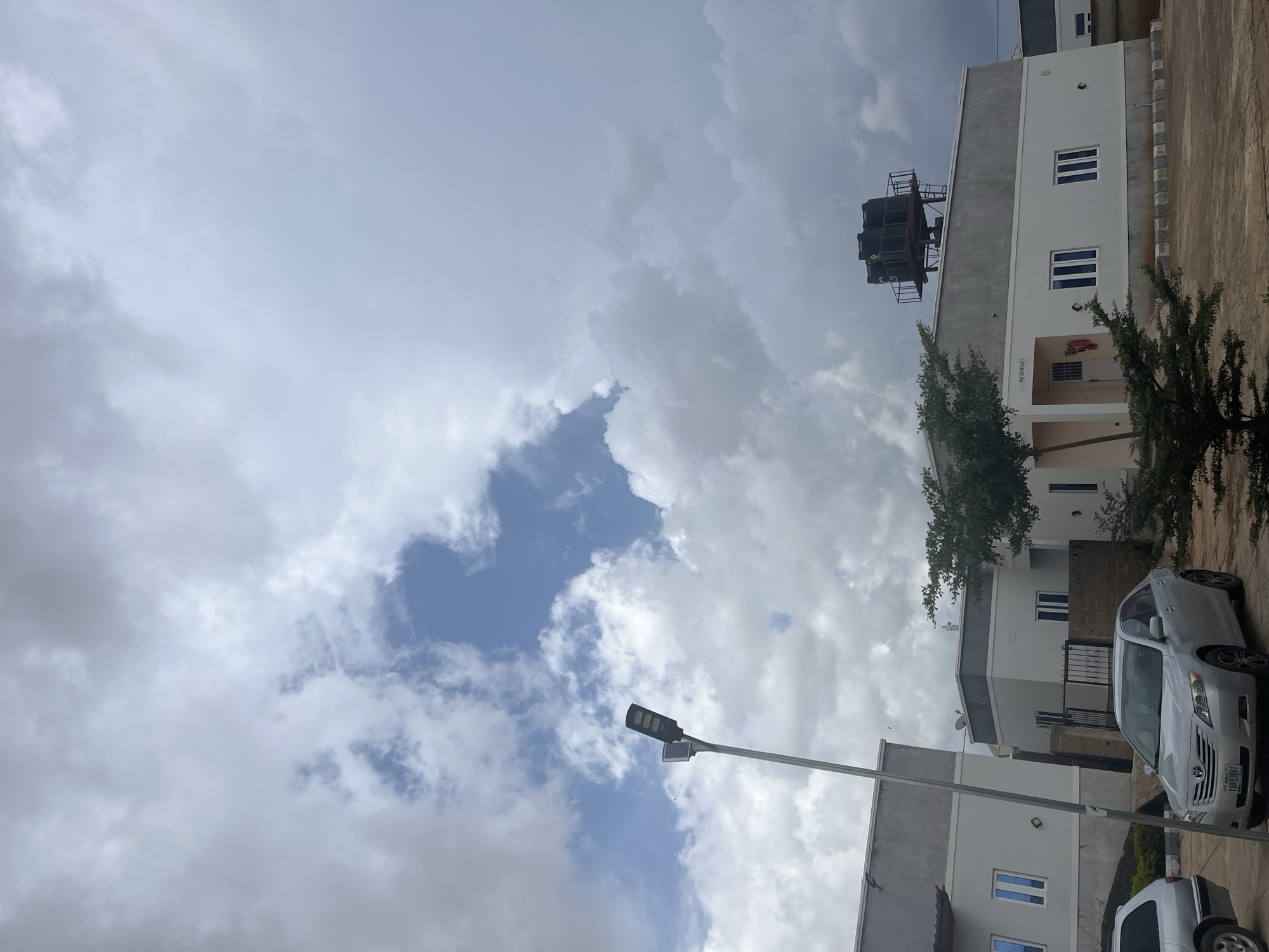 The image shows a row of modern, light-colored buildings with grey accents, likely a commercial or residential complex, situated in Jos, Nigeria. The sky is overcast with dramatic, textured clouds, suggesting a change in weather. In the foreground, a silver Toyota Camry is parked on a paved surface. To the left, part of a silver SUV is visible. A tall, dark lamppost with a solar panel is positioned prominently in the left half of the frame, casting a long shadow. The buildings feature multiple windows and what appears to be a fire extinguisher and a sign for "PHYSIOTHERAPY" on one of them. A metal structure supporting several dark water tanks is visible on the roof of one of the buildings. A small, sparsely leaved tree stands in front of the physiotherapy center. The overall impression is of a well-maintained, contemporary development in an outdoor setting.