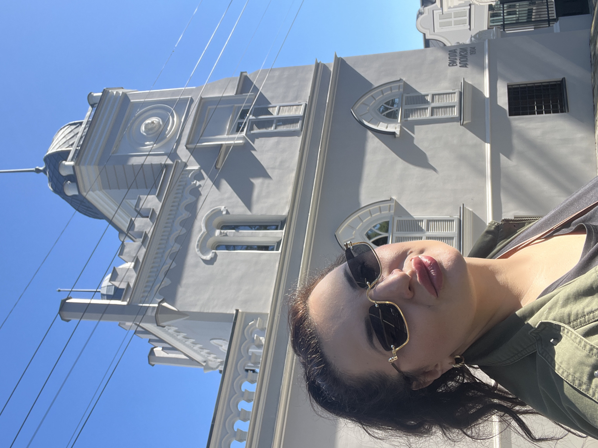 A portrait-style photograph captures a woman posing in front of a ornate building on a bright, sunny day. The image is taken at an angle, with the woman's face and upper body dominating the foreground on the right, and the building extending behind and to the left. The woman has dark hair and is wearing sunglasses with gold frames. Her lips are full and tinted with what appears to be a reddish-brown lipstick. She is wearing a dark top under an olive-green jacket.
The building behind her is painted a light, possibly off-white or light gray color, featuring elaborate architectural details. It has multiple windows of various shapes, including arched and rectangular ones, some with shutters. A prominent feature is a dome or cupola visible on the far left of the building's roofline. Decorative cornices, balustrades, and reliefs are visible along the structure. The clear blue sky fills the background above the building. Several thin, dark lines, possibly power lines, stretch across the sky in the upper left portion of the image.
On a section of the building's facade, text is visible: "ΒΙΛΛΑ ΑΛΟΗ" (which translates to "Villa Aloi" or "Villa Aloe") and below it, the year "1904." The sun casts sharp shadows from the building's features, indicating strong sunlight.
