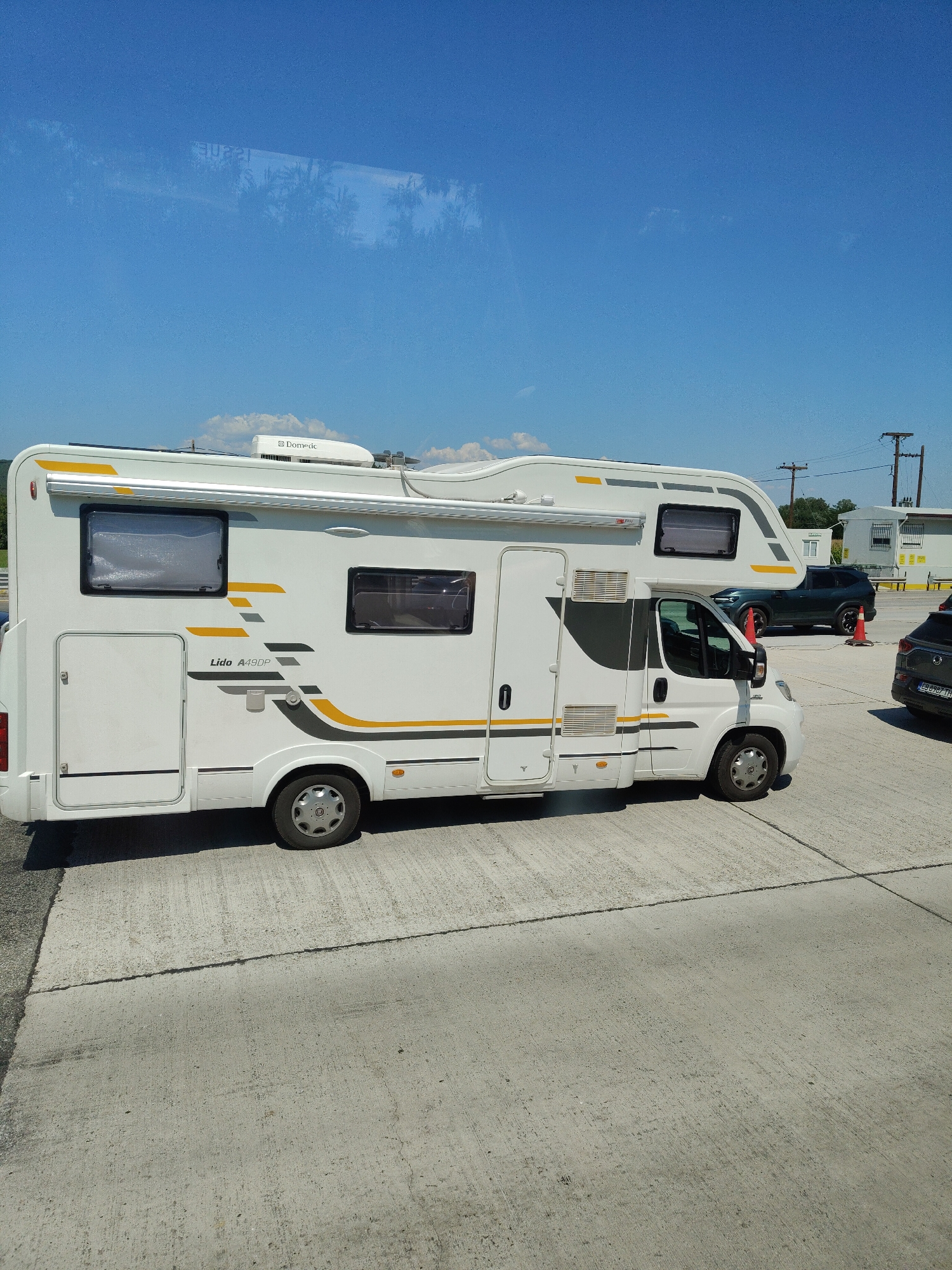 A white camper van with yellow and grey accents is parked on a concrete surface under a clear blue sky. The camper van has multiple windows, an awning, and a Dometic air conditioning unit on the roof. In the background, a dark green SUV is parked behind an orange traffic cone, and another dark-colored vehicle is visible to the right. There are power lines and small buildings in the distance. The scene appears to be a parking lot or roadside area in the Municipality of Promachonas, Greece.