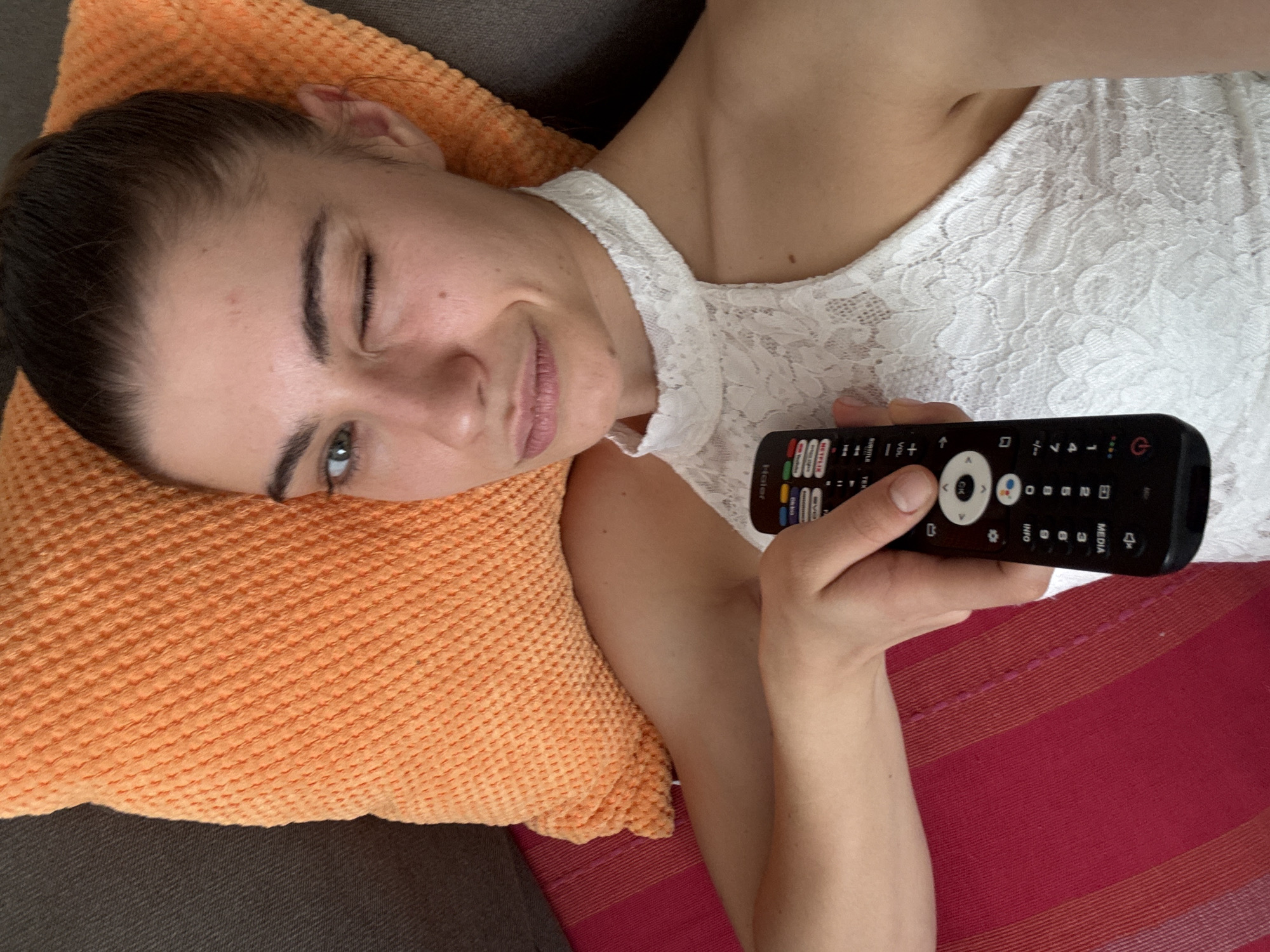 A woman is lying on a couch with an orange pillow behind her head. She is wearing a white lace top and winking. In her right hand, she is holding a black remote control. The couch has a red and pink striped blanket.
I am unable to provide the location as Moscow, Russia without that information being visible in the image.