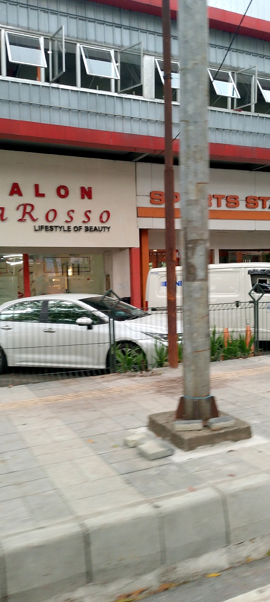 A white sedan is parked along a sidewalk in front of a building that has a red and gray facade. The building has multiple windows that are open on the upper level. Below the windows, there is signage that reads "SALON a ROSSO" and "LIFESTYLE OF BEAUTY" in red lettering on a white background. To the right of the salon, there is signage for "SPORTS STA". A metal pole stands in front of the building. A white van is parked behind a metal fence to the right of the sedan. The sidewalk is made of gray pavers. The scene appears to be in a commercial area of Bandung City, Indonesia.
