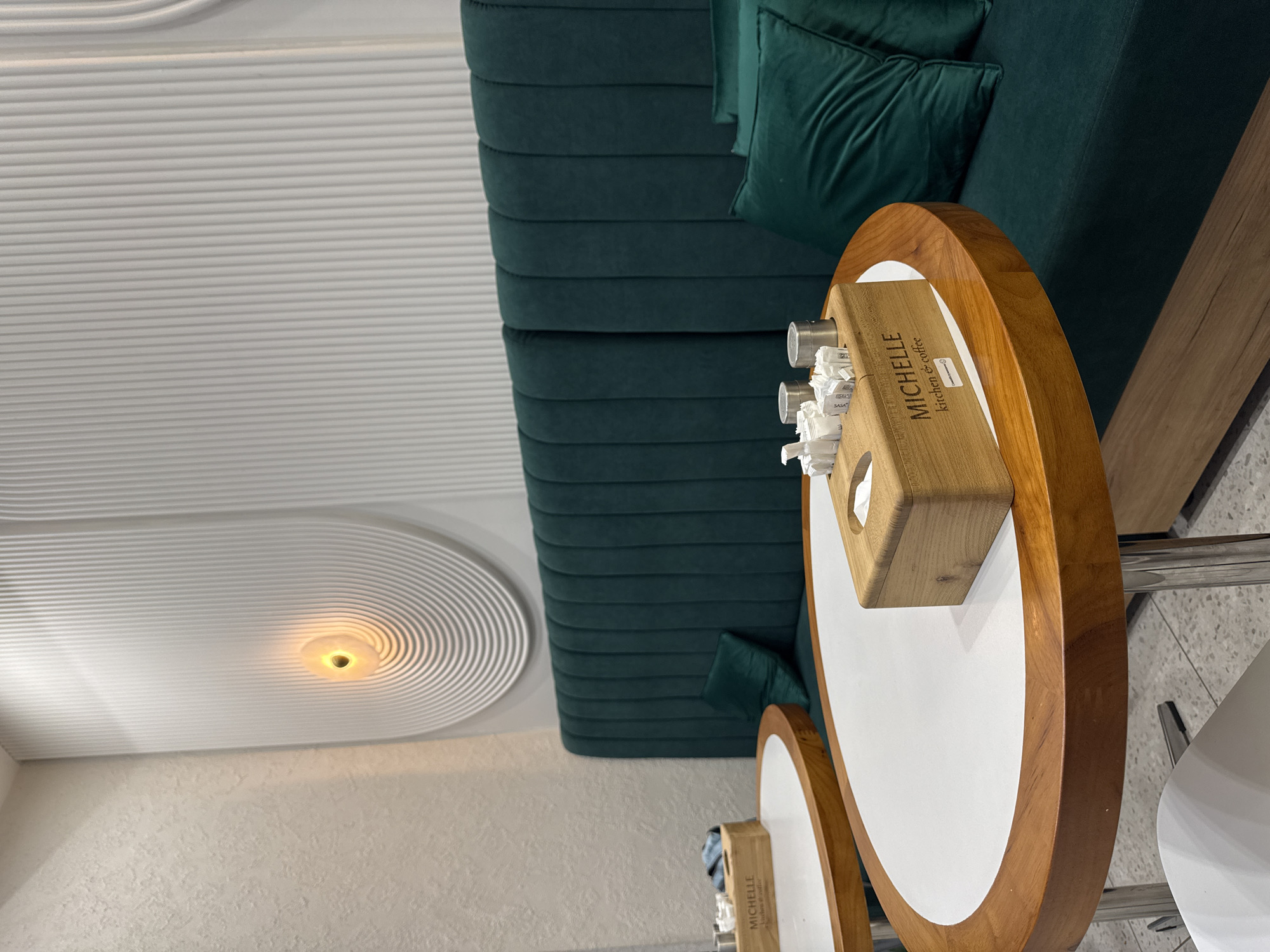 The image shows an interior view of a modern cafe, possibly in Bishkek City, Kyrgyzstan. The cafe features a deep teal upholstered banquette seating area against a white wall with textured finishes and a decorative ribbed paneling design. A circular, recessed light fixture with a warm glow is embedded in the ribbed panel.
In the foreground, two oval-shaped tables with white tops and wooden rims are visible. One table has a wooden organizer filled with sugar packets, stirrers, and napkins. The organizer has the text "MICHELLE kitchen & coffee" printed on it. A second table to the left also has a wooden organizer with napkins. The tables are supported by chrome legs.
There are two plush teal cushions on the banquette. The floor appears to be tiled with a speckled pattern. The overall atmosphere is clean and contemporary, suggesting a comfortable dining or coffee-drinking environment. No people are present in the image.