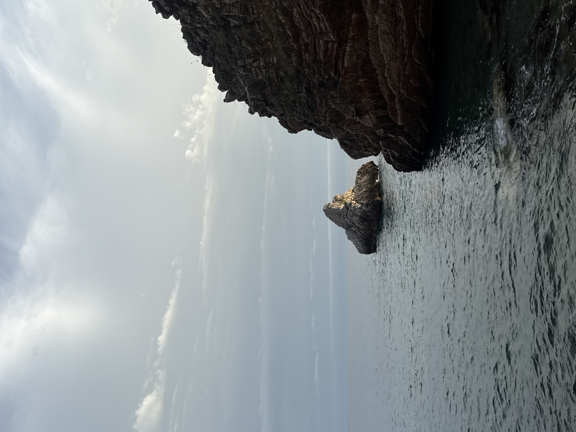 The image depicts a coastal scene with a rugged rock formation dominating the right side of the frame. The rock is dark brown and shows layered geological strata, with some vegetation growing on its upper surfaces. In the distance, a smaller, pointed rock formation rises from the calm, dark blue water. The sky is overcast with light gray and white clouds, and a subtle sunlight illuminates the peak of the distant rock. The water is gently rippling, creating a textured surface. The perspective suggests a view from the shore or a boat near the coast, facing the sea. The location is identified as the city of Rafailovići, Montenegro.