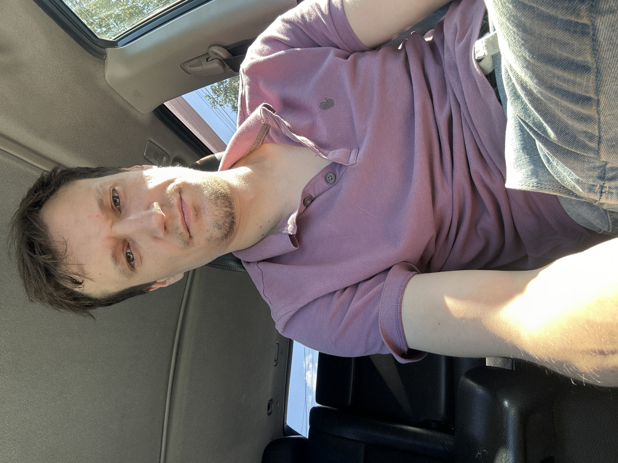 The image shows a man sitting in the driver's seat of a vehicle. He is looking directly at the camera, with his head tilted slightly to his left. He has short, dark brown hair and is clean-shaven except for a light stubble on his chin and upper lip. He is wearing a pink polo shirt with two buttons at the collar, and the collar is popped up on the right side. The interior of the vehicle is visible, including the dashboard, the passenger seat, and part of the back seat. The windows are open, and trees and a blue sky are visible outside. The image appears to have been taken during the daytime, with sunlight casting shadows on the man's face and arm. There are no specific landmarks or context provided to indicate a location within the city of Makiv, Ukraine.