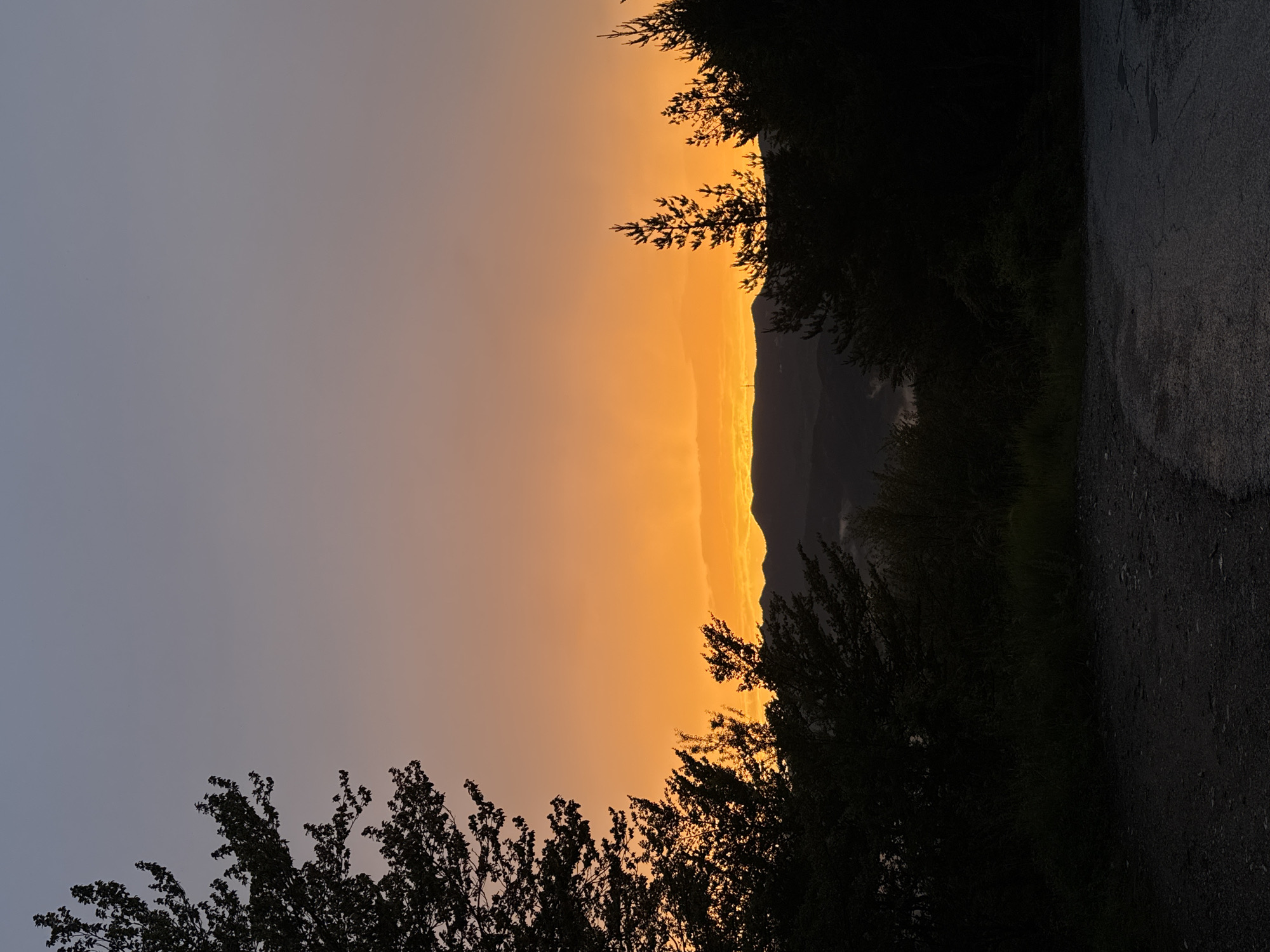 The image depicts a serene natural landscape at dusk in Strazha, Bulgaria. Silhouetted trees frame the foreground, with a dirt road leading into the scene. Beyond the trees, rolling hills are visible against a gradient sky that transitions from a deep blue at the top to a vibrant orange and yellow near the horizon, indicating the setting sun. The overall atmosphere is tranquil and picturesque.