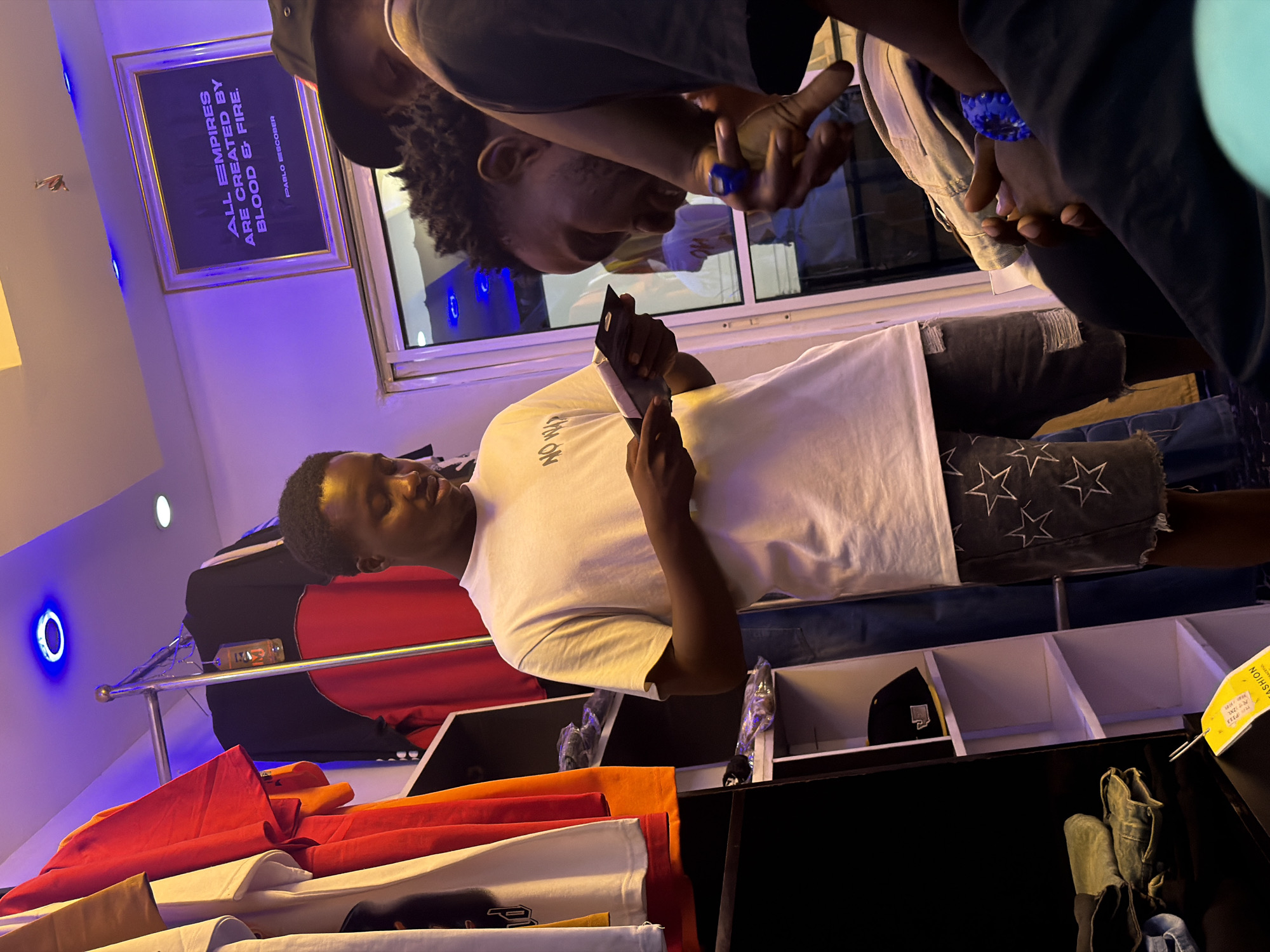 In a retail setting in Bukuru, Nigeria, three young Black men are present. One individual in the foreground, facing slightly right, wears a white t-shirt with "NO WAY" printed on it and denim shorts adorned with white stars and distressed details. He holds a small booklet or pamphlet. To his right, another man, viewed from the side, appears to be looking down and smiling, his hair styled in short, curly locs. He wears a dark t-shirt and has a blue ring on his right index finger. Partially visible in the background, a third man wears a black hat and a dark t-shirt. The environment is brightly lit with purple and blue LED lighting. Clothing racks with various t-shirts are visible on the left, and shelves with apparel and a baseball cap are on the right. A framed picture with text reading "ALL EMPIRES ARE CREATED BY BLOOD & FIRE. PABLO ESCOBAR" is displayed on the wall.