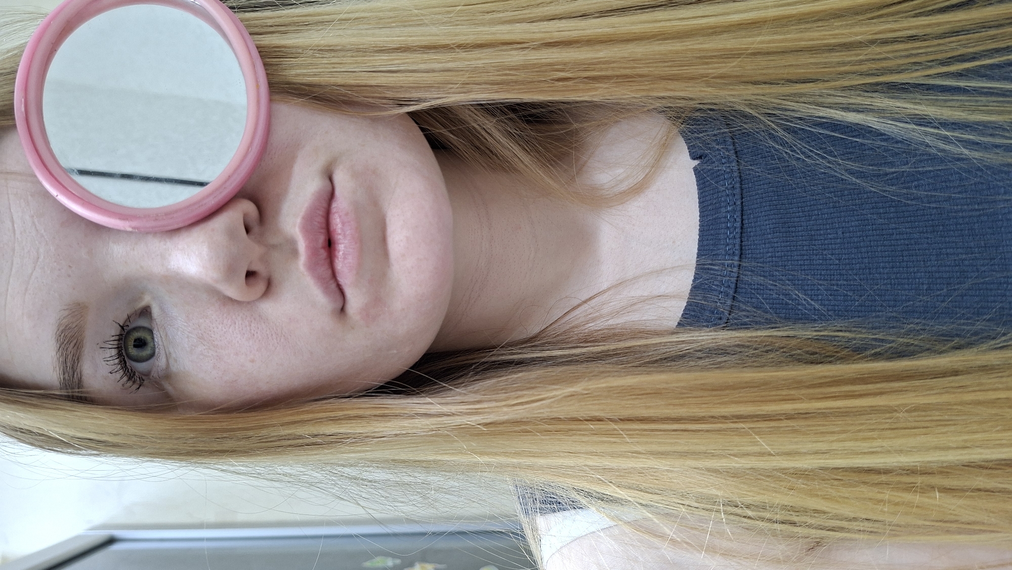 A young woman with light skin, fair hair, and green eyes is lying down. She is wearing a blue ribbed tank top. A pink-framed round mirror is positioned over her left eye. The setting is indoors and the location in the city of Большой Букор, Russia cannot be determined from this image.