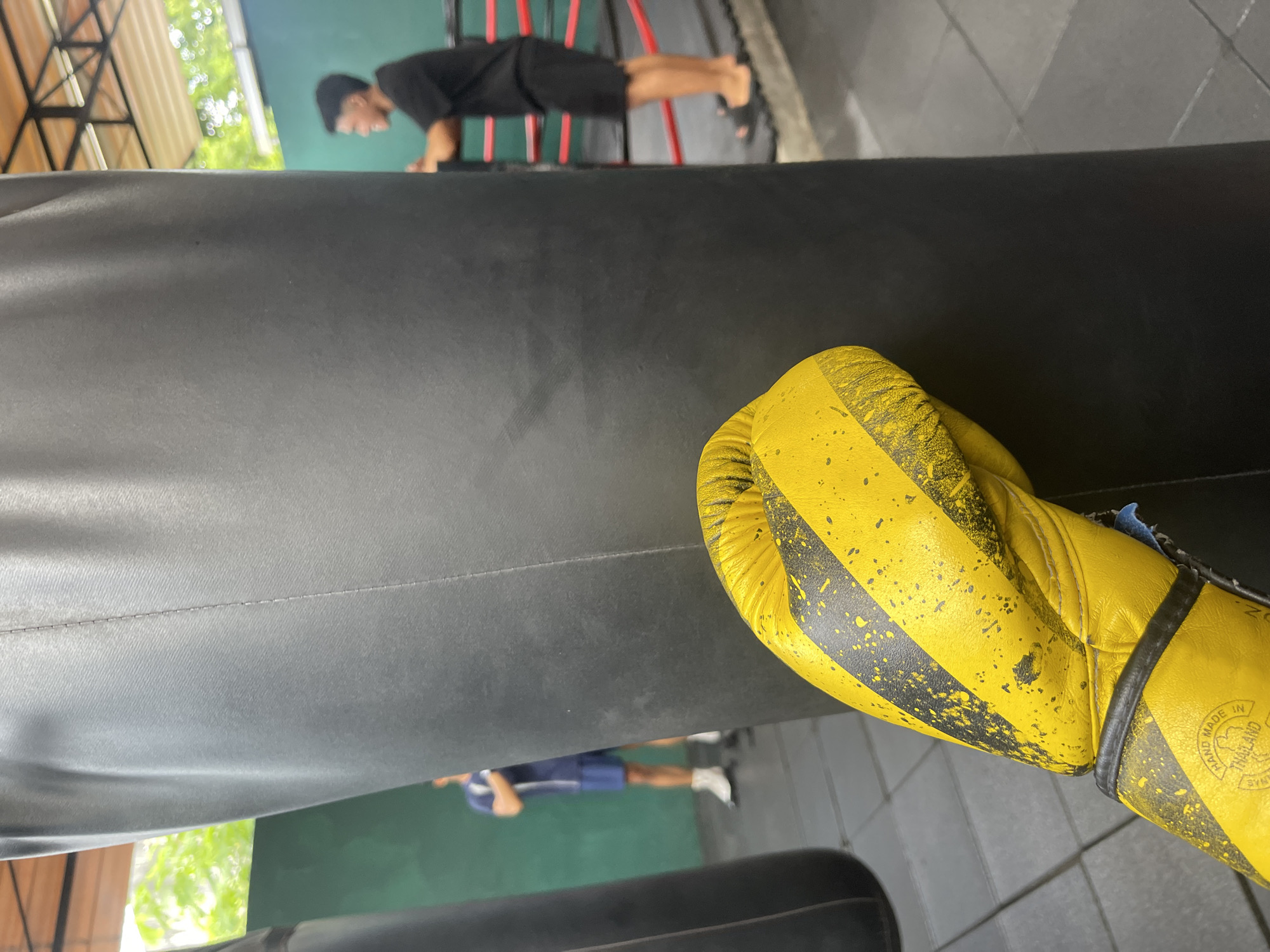 In Kuta Selatan, Indonesia, a close-up shot focuses on a yellow boxing glove with black stripes, resting against a large black punching bag. The glove has visible wear and tear, with speckles of black paint or scuffs. In the background, slightly out of focus, two individuals are present. One person, wearing a dark t-shirt and shorts, stands in a boxing ring with red ropes. Another person, in the middle ground and wearing a blue t-shirt and shorts with white socks, stands near another punching bag. The environment appears to be an indoor gym or training facility, with a tiled floor and a partially visible open-air ceiling structure.