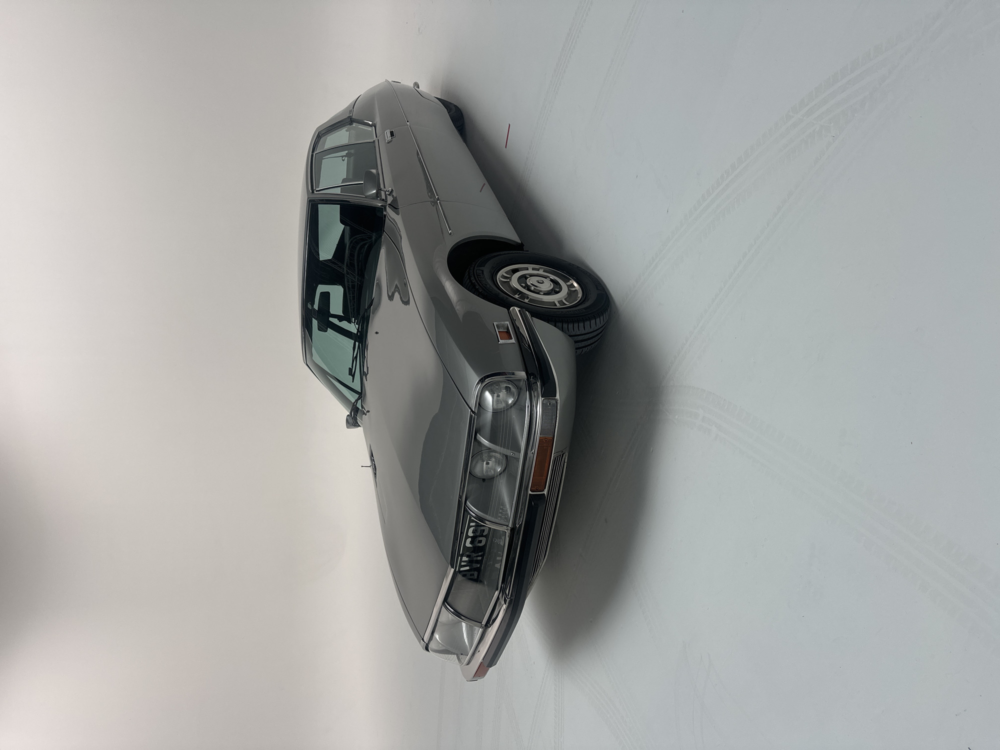 A light grey or silver vintage Citroën SM car is prominently displayed in an indoor studio environment. The vehicle is positioned facing left, captured from a slightly elevated, front-quarter perspective. Its distinctive front end features a unique glass cover encasing multiple headlights, with an orange indicator light visible on the right side. The front license plate clearly reads "AWR 69J." The car's classic design includes intricate wheel covers and a sleek body, appearing to be in pristine, well-maintained condition.
The setting is a minimalist studio with a seamless light grey or white backdrop and floor, suggesting professional photography or display. Subtle tire track impressions are visible on the studio floor, especially towards the right side of the image, indicating past movement or positioning of vehicles. The lighting is even and bright, typical of a controlled studio setup, highlighting the car's contours without harsh shadows. No people are present, and the car is stationary. The British license plate "AWR 69J" corroborates the provided location context of London, United Kingdom.