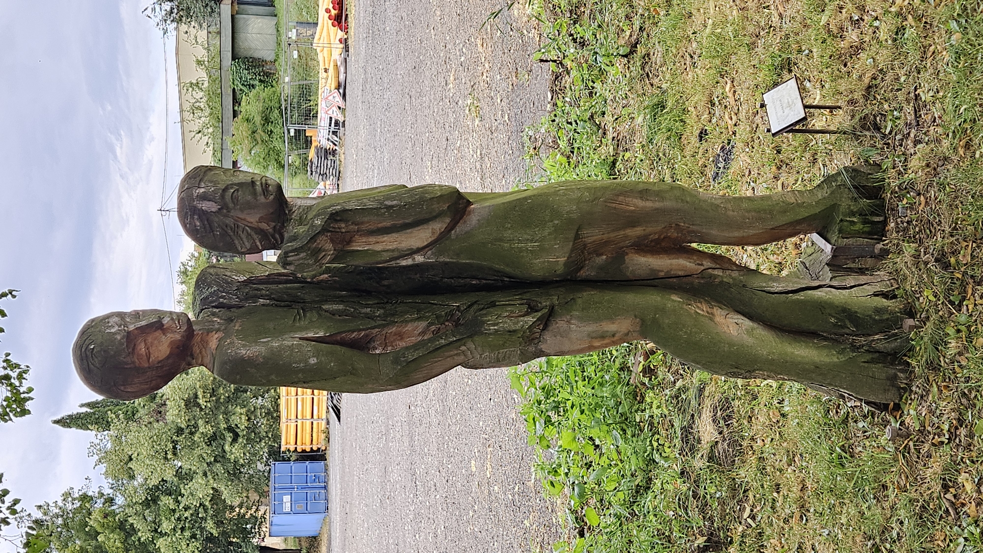The image shows a wooden sculpture of two intertwined figures, possibly representing a couple or a close bond. The sculpture is weathered and covered in moss, suggesting it is outdoors and has been exposed to the elements for some time. The figures appear to be embracing or leaning into each other, conveying a sense of connection or intimacy. Their faces are carved with simple, somewhat abstract features. The sculpture is positioned vertically, with its base resting on the ground.
The setting appears to be a park or a public outdoor space. To the left of the sculpture, there are trees and foliage, indicating a natural environment. In the background, there are hints of urban elements: a building with a light-colored facade, a chain-link fence, and what looks like industrial equipment, including yellow structures and a blue shipping container. A paved path runs alongside the sculpture. The ground surrounding the sculpture is covered with grass, fallen leaves, and some small plants.
A small, white sign with some text is visible on the right side of the sculpture, planted in the grass. The text on the sign is illegible in the provided image.
The lighting suggests it is daytime, likely a bright day with some clouds in the sky. The overall mood conveyed by the sculpture is one of quiet contemplation and enduring connection. The location cues suggest this might be in an area that blends nature with industrial or urban infrastructure. Given the provided location context of Prešov, Slovakia, this sculpture is likely a public artwork situated within the city or its surroundings.