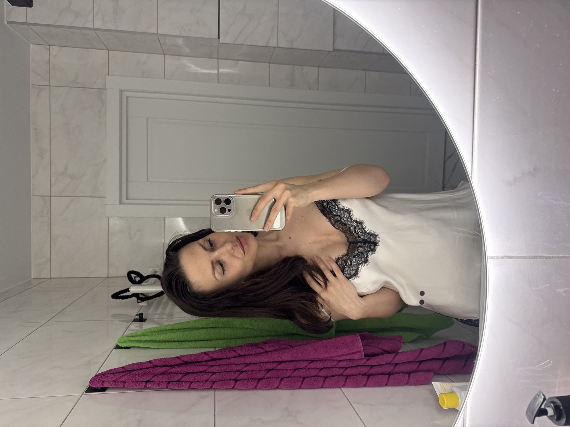 The image depicts a woman taking a selfie in a bathroom. She is holding a smartphone in her right hand, partially obscuring her face. She has long, dark hair. She is wearing a white top with black lace trim around the neckline.
The bathroom is tiled with white rectangular tiles. A mirrored cabinet with white doors is visible in the background.
Two towels are lying on the floor: one green and one purple.
A black hairband and a tube of cream are also visible on the floor.
There is no discernible information to determine if the location is in Murino, Russia.