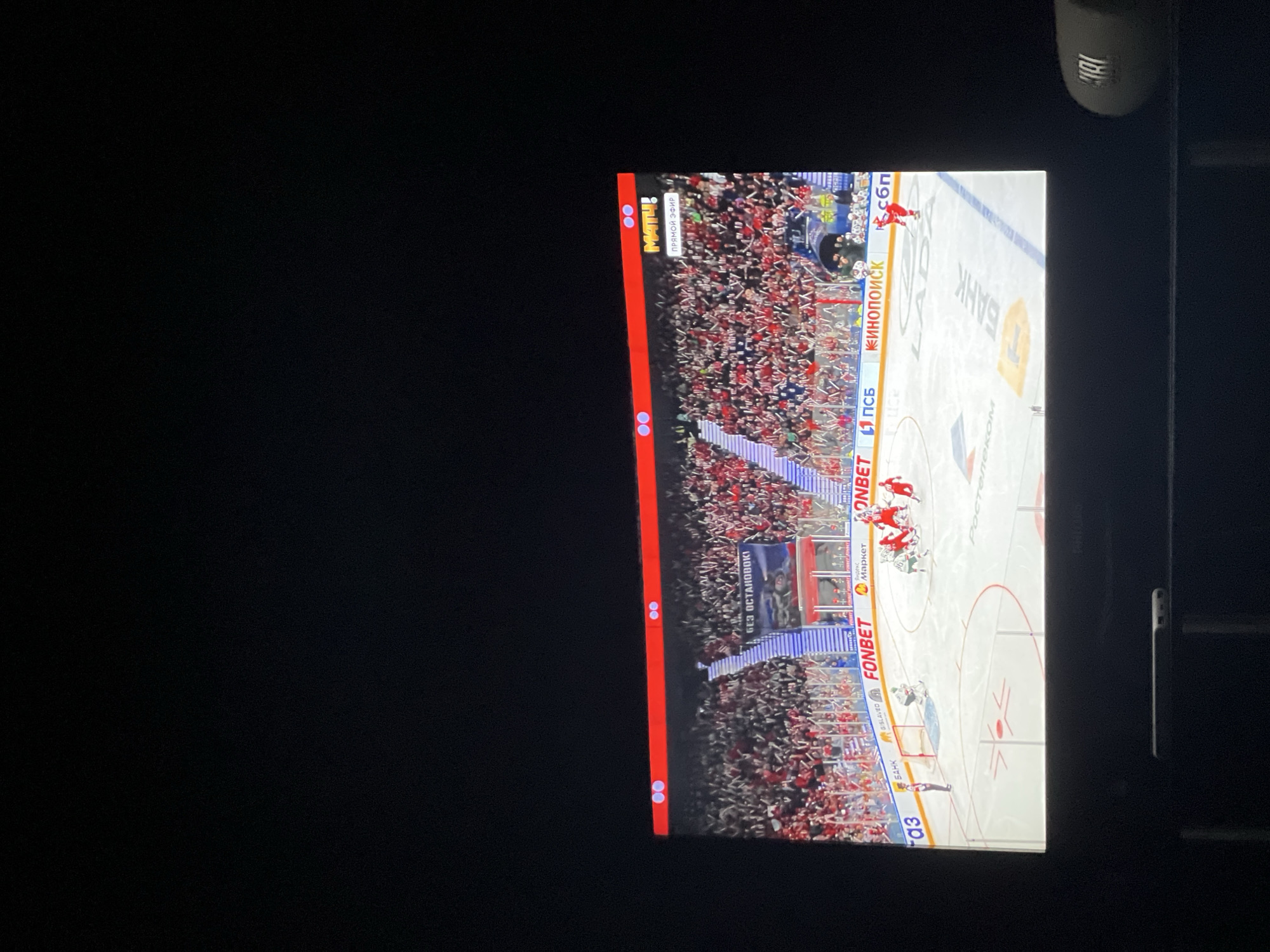 The image shows a TV screen displaying a hockey game. The ice rink is visible with players in red and white jerseys. Crowd in stands around rink. Text on rink "FONBET", "БАНК", "КИНОПОИСК". Upper left corner of the screen, the television channel is "MATH!". The game is "ПРЯМОЙ ЭФИР" ("Live Broadcast"). The environment around the TV is dark. There is a JBL speaker in the upper right corner of the image. Based on the Russian text, it is likely a game in Russia, possibly in the city of Krasnoyarsk, Russia, given the text "БЕЗ ОСТАНОВOKI" (no stops).