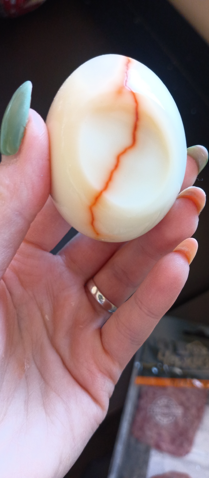 A hand with long nails, some painted green and some orange, holds a peeled hard-boiled egg with a red vein-like mark running across its surface. A silver ring is visible on the ring finger of the hand. Part of a package with blurry, red contents is visible in the lower right corner of the frame. The background is dark. The location is Unknown, Unknown.
