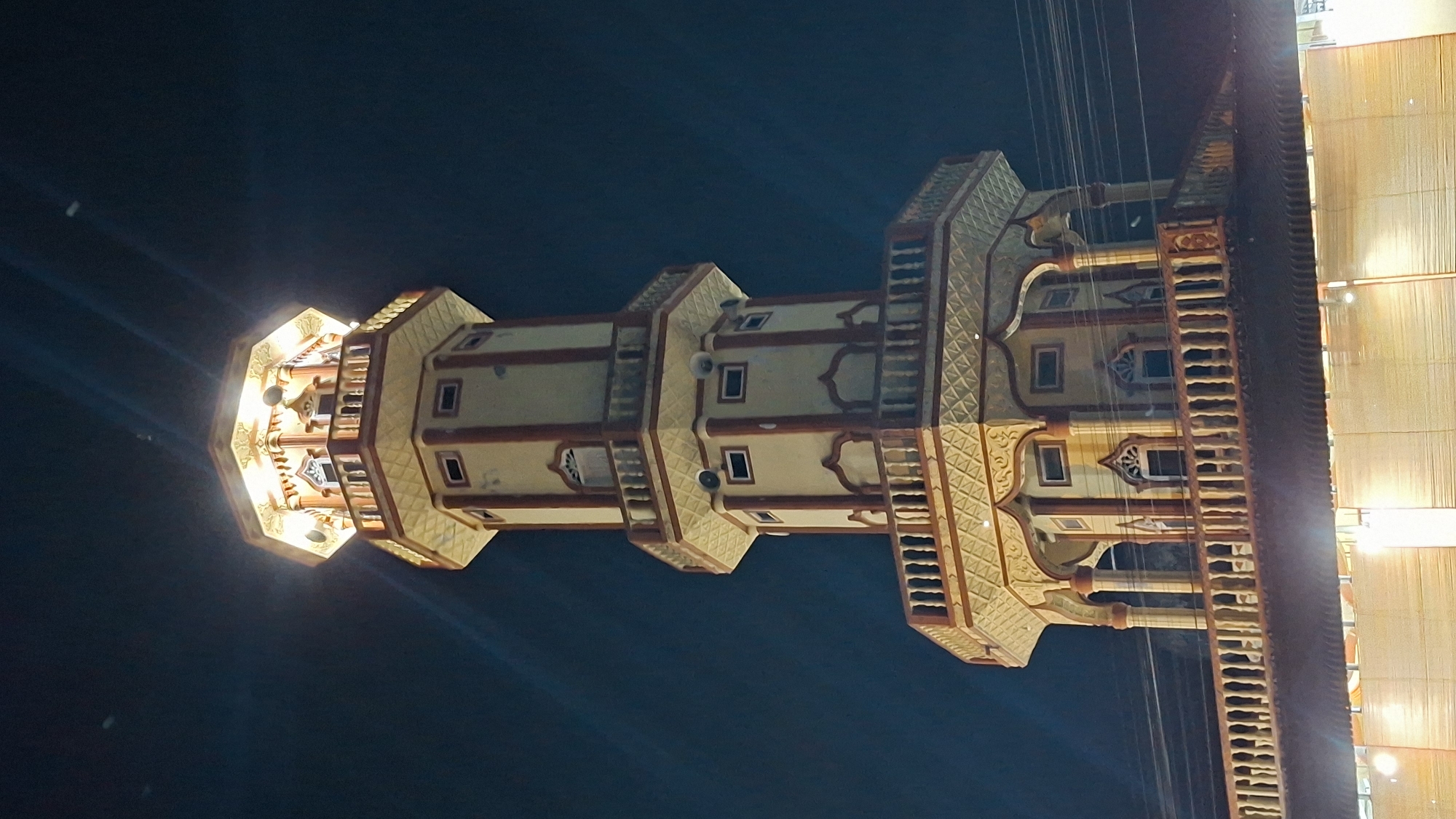 A multi-tiered tower, illuminated at night, stands against a dark sky. The tower has an octagonal base and tapers upwards, featuring balconies with ornate railings and arched windows. The top section is brightly lit, revealing decorative elements. Power lines are visible in the background, and a dimly lit structure with a thatched roof is in the foreground. The image is taken from a low angle, looking up at the tower. The location is identified as Keudeteupinpunti, Indonesia.