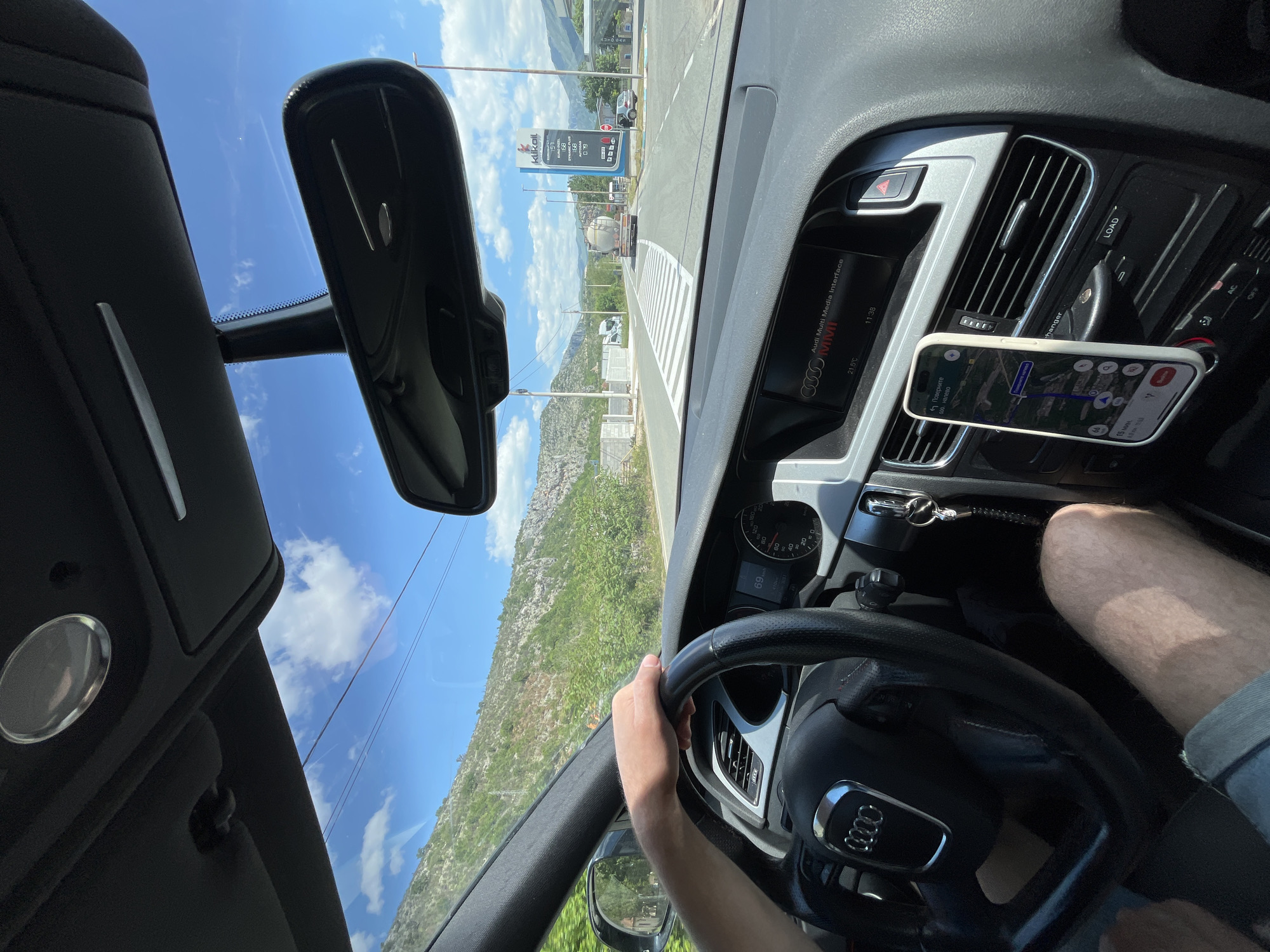 A first-person shot from the driver's seat of an Audi car. The driver's left hand is on the steering wheel. The dashboard contains the Audi Multi Media Interface (MMI) display showing "21.5°C" and "11:38," next to it is a cell phone with a navigation app displaying a turn-by-turn route. Through the windshield, a rural road bordered by green shrubs and mountains is visible. A "klikoil" sign stands roadside. The scene is located in Donje Mrke, Montenegro.