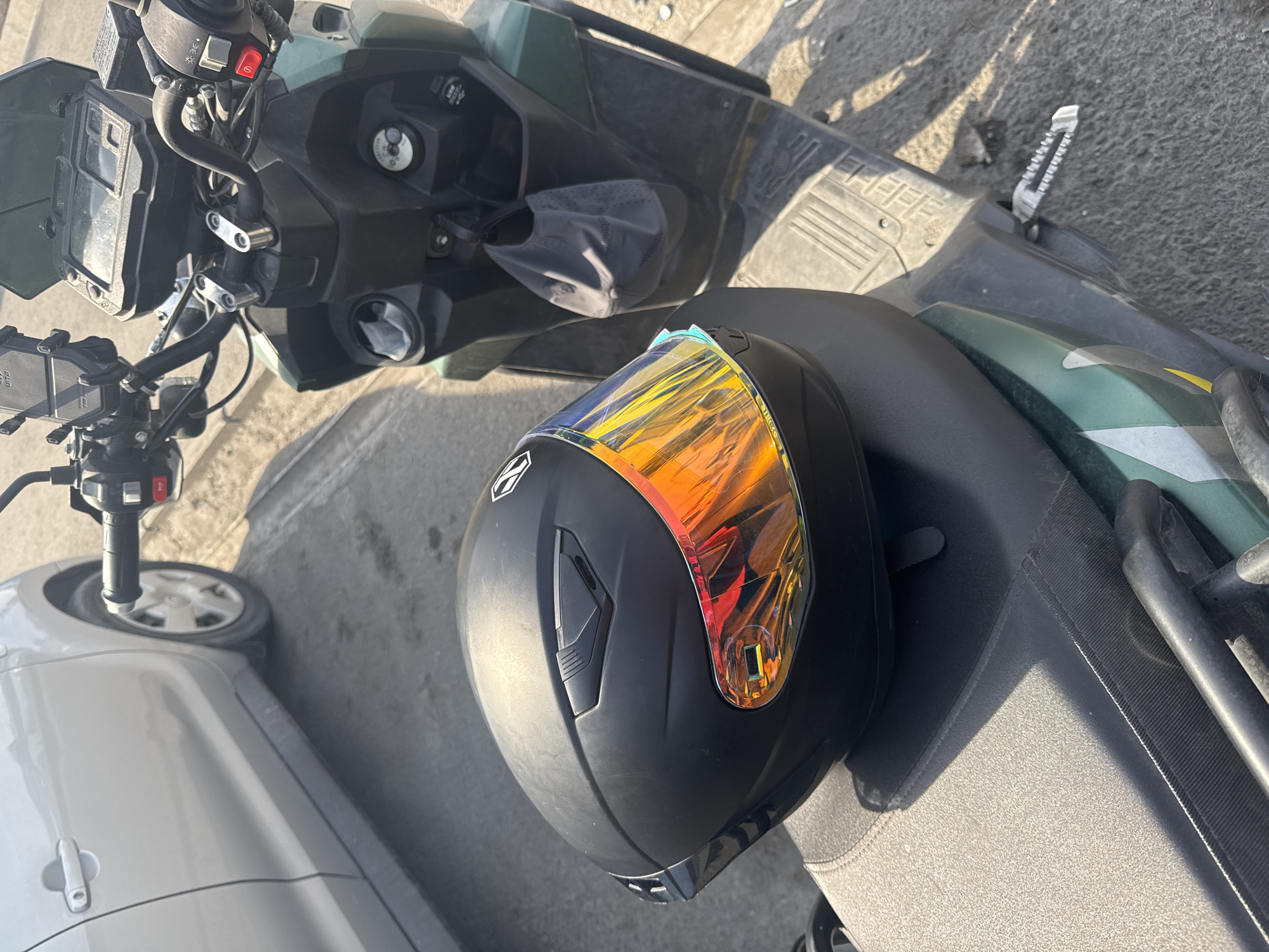 In Bishkek City, Kyrgyzstan, a black motorcycle helmet with a reflective, iridescent visor rests on the dark-colored seat of a dark green scooter. The scooter's handlebars, instrument panel, and front portion are visible. To the left of the scooter, the side of a silver car is parked on a paved surface, with its rear tire and lower body in view. The scene is bathed in sunlight, casting shadows on the ground.