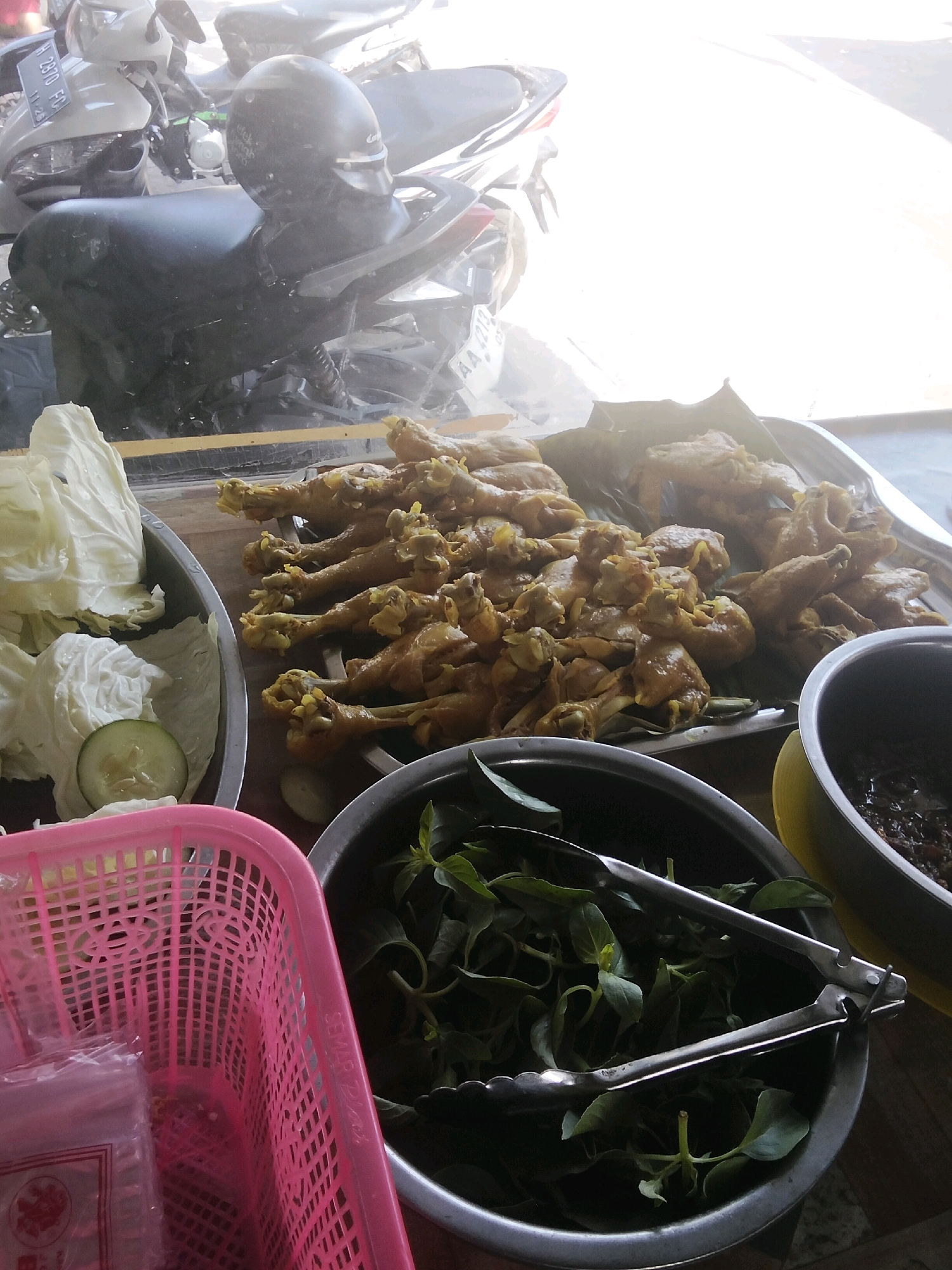 The image captures an outdoor food stall in Magelang, Indonesia, set against the backdrop of parked motorcycles and a bright, hazy sky. In the foreground, a pink plastic basket holds what appear to be wrapped utensils or food packets. To the right of the basket, a stainless steel bowl overflows with fresh green leaves, likely herbs or vegetables, with a pair of metal tongs resting inside.
Further back, a large metal tray displays a generous portion of fried chicken pieces, some arranged on a banana leaf. A bowl of dark sauce or relish is visible to the right of the tray. To the left, a large metal bowl contains chopped cabbage and a slice of cucumber, with a hint of what looks like white cream. The scene is illuminated by strong sunlight, creating a somewhat washed-out effect and casting soft shadows. The overall impression is of a humble, ready-to-serve food offering in a busy urban environment.