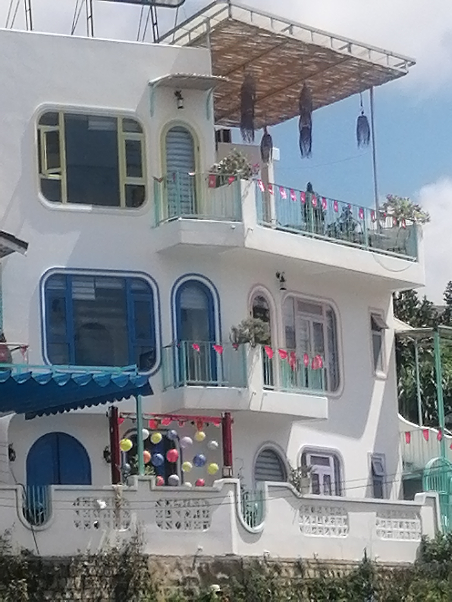 The image depicts a multi-story white building with distinctive curved architectural features and rounded windows. The building is adorned with festive decorations, including colorful balloons and small flags. The upper portion of the building is partially covered by a bamboo-like thatched roof structure. The scene is set against a blue sky with some clouds, and lush green foliage is visible in the foreground and background. This building appears to be a cafe or guesthouse in Da Lat, Vietnam, known for its unique architecture and charming ambiance, often decorated for holidays or special events.