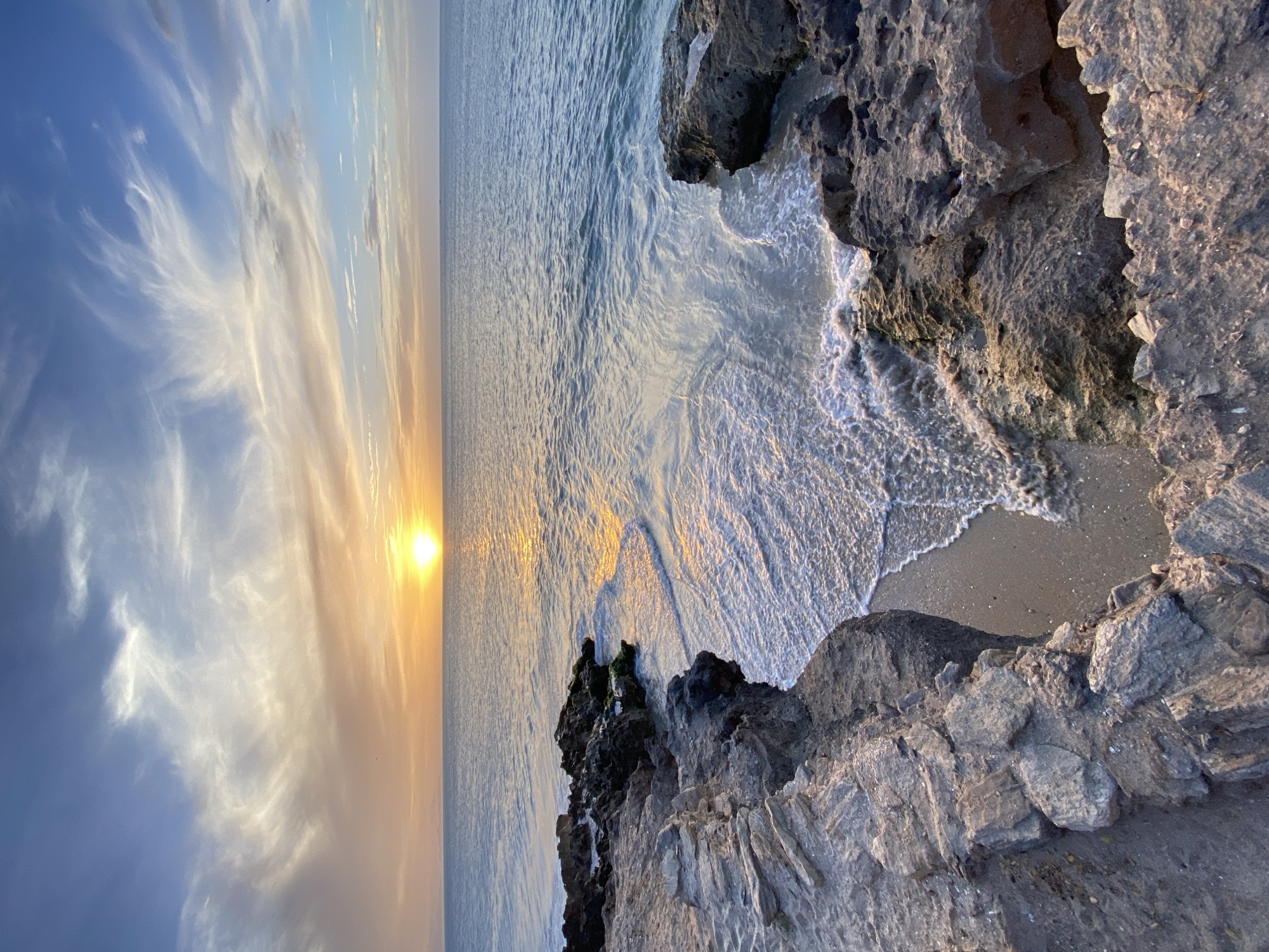 A vertical shot captures a serene seascape during sunset. The sky, occupying the left half, transitions from blue to hues of orange and yellow near the horizon, showcasing wispy clouds. A radiant sun sits on the horizon, its light reflecting on the water, creating a golden path stretching towards the viewer. The right half features rugged, light-colored rocks forming a cliff edge, with gentle waves washing onto a small sandy beach below. This image is likely from Juan Griego, Venezuela. There are no people visible in the shot.
