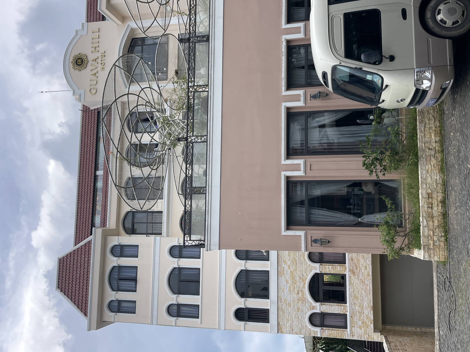 The image shows the exterior of the Guava Hill Hotel in Lâm Viên - Đà Lạt, Vietnam, on a cloudy day. The hotel has a light beige facade with a red tiled roof. The building features multiple arched windows with dark frames and some with ornamental grilles. A prominent sign at the top of the building reads "GUAVA HILL HOTEL" in gold lettering, set within an oval emblem. There is a decorative metal structure on a balcony overlooking the street. In the foreground, a white and beige passenger van is parked on a cobblestone driveway. The lower part of the hotel is clad in textured beige stone. A garage door is visible to the left of the van. Greenery, including small bushes and trees, is present around the base of the building.