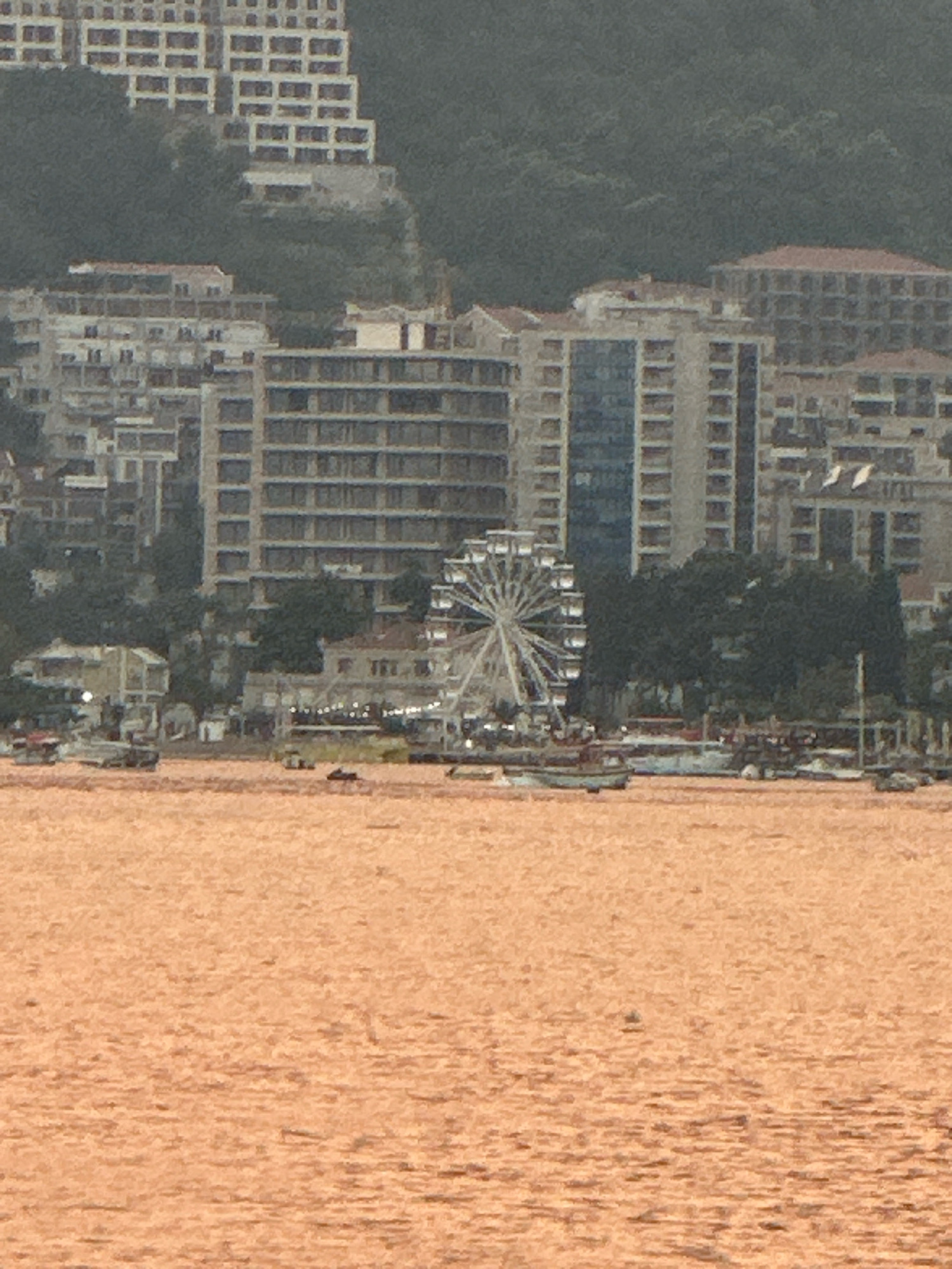 The image shows a panoramic view of the Budva coastline in Montenegro. In the foreground, a body of water, possibly the Adriatic Sea, reflects the light with an orange hue. Scattered on the water are several small boats, some appearing to be moored. In the middle ground, a Ferris wheel stands prominently against a backdrop of modern buildings and hotels that cascade down a hillside. Trees and vegetation are visible between the buildings. The buildings themselves exhibit a mix of architectural styles, with some featuring balconies and many windows. The hillside behind the buildings is covered in dense, dark green foliage. The sky above is overcast, casting a soft, diffused light on the scene.