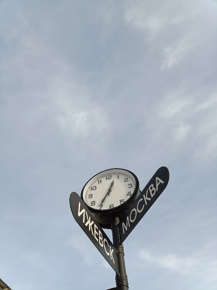 The image displays a street clock and two directional signs affixed to a dark vertical pole, set against a light gray, partially cloudy sky. The central element is a round, white-faced analog clock featuring black Arabic numerals from 1 to 12 and black hour and minute hands, indicating approximately 1:35. The clock face is slightly tilted upwards and to the viewer's right. Below the clock, two black, elongated directional signs extend from the pole. The sign angled towards the lower-left displays "ИЖЕВСК" (Izhevsk) in white Cyrillic letters. The sign angled towards the upper-right displays "МОСКВА" (Moscow) in white Cyrillic letters. The sky occupies the majority of the frame, with only a very minimal, unidentifiable structure faintly visible at the bottom left edge. This scene is located in a public space within the city of Chaikovsky, Russia.