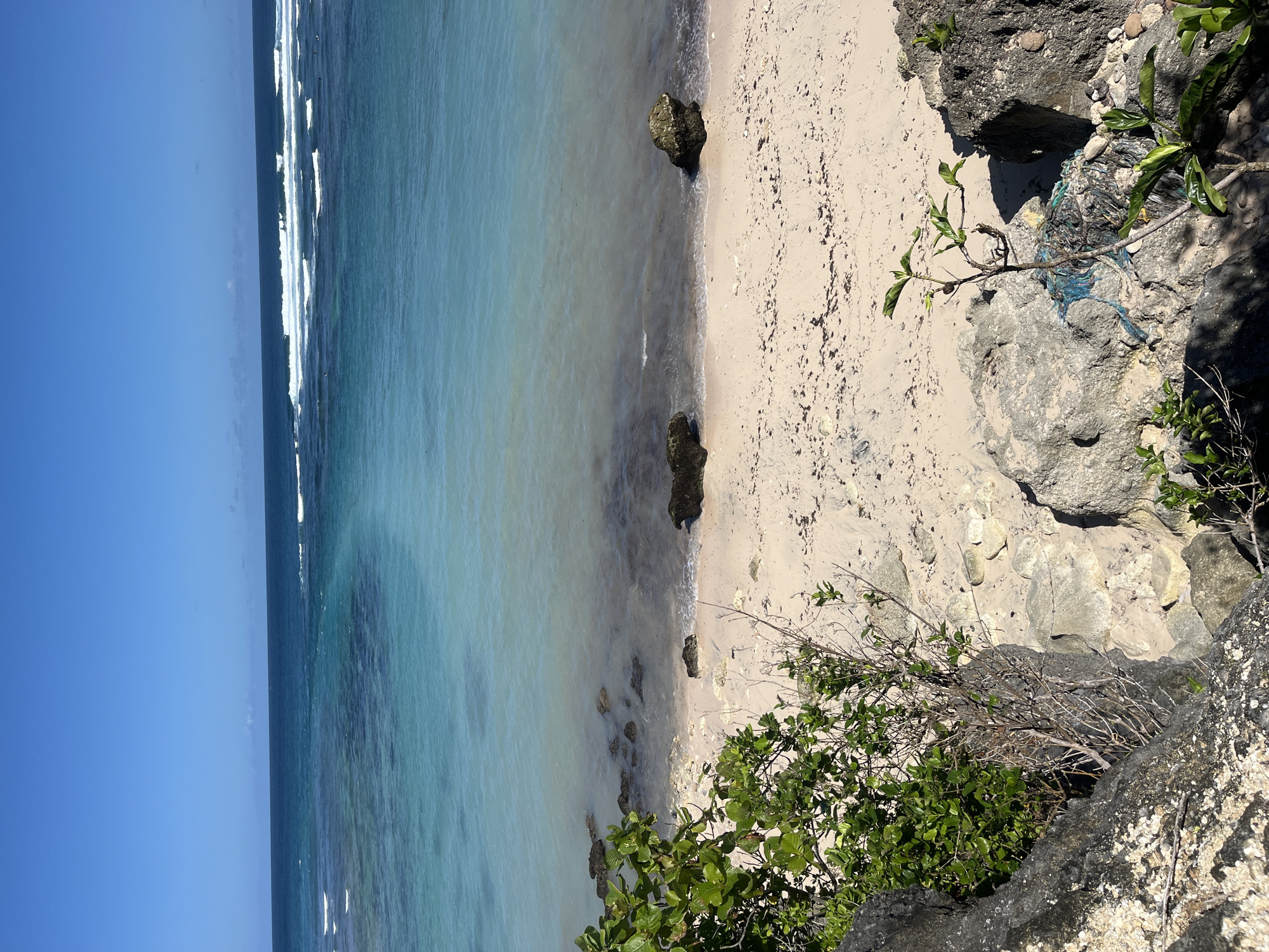 The image depicts a beach scene from an elevated viewpoint, likely along the coastline. The location appears to be Kuta Selatan, Indonesia. The scene features a narrow strip of light-colored sand along the coastline, adjacent to a body of clear turquoise water. The water transitions from lighter shades near the shore to darker hues further out. Small waves can be seen along the edge of the ocean. On the right side of the image, a rocky cliff formation covered with shrubs is visible. A small tree branch is seen near the rocks. The sky is clear and blue. The photograph appears to be taken during daylight hours. There are no people visible in the scene.