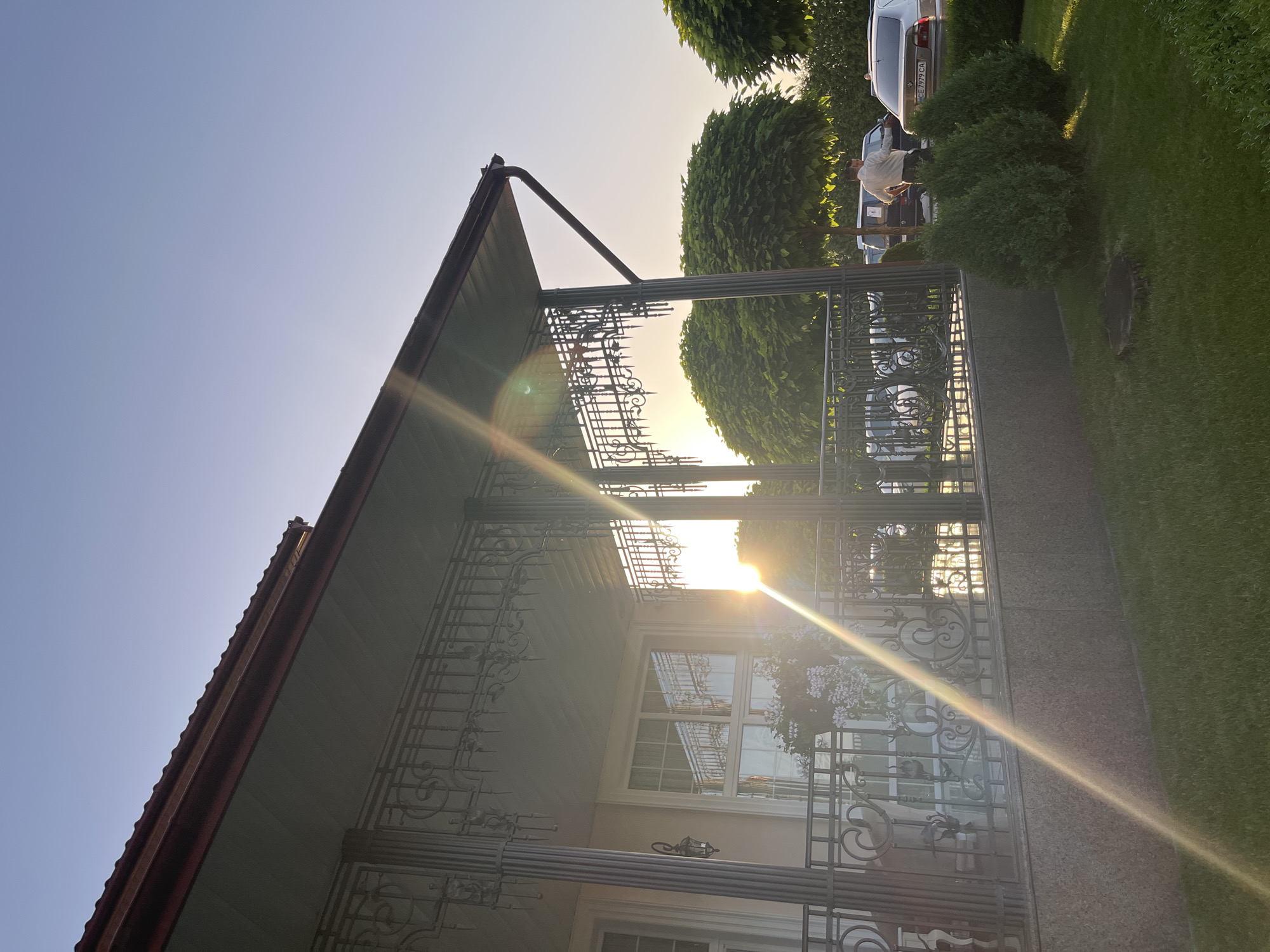 The image depicts a sunlit scene in Dubivtsi, Ukraine, likely during the late afternoon or early evening due to the low angle of the sun and the warm, golden light casting long lens flares across the frame. The photograph is taken from a low perspective, looking upwards towards the roof of a house and the surrounding exterior.

The main subjects include the architecture of a house with a dark-tiled roof and a green soffit, partially obscured by decorative metal railings that form a porch or balcony. These railings are intricately designed with scrollwork and finials. Beyond the railings, a large window with a grid pattern is visible, reflecting the bright sunlight. A hanging basket with delicate pink flowers adds a touch of color to the scene.

In the background, several lush green trees with rounded canopies are prominent, framing the sky. To the right, a patch of well-maintained green grass and a paved pathway are visible. Further back, a silver car is parked, and a person, dressed in a light-colored shirt, appears to be interacting with the car or the surrounding bushes. The overall setting suggests a residential area with well-kept landscaping.

The bright sunlight and the lens flares create a vibrant and somewhat hazy atmosphere. The clear blue sky indicates fair weather. The time of day is suggested by the direction and intensity of the sunlight, which appears to be setting or low in the sky. The location context of Dubivtsi, Ukraine, provides a geographical anchor for the scene. No visible text is present in the image.