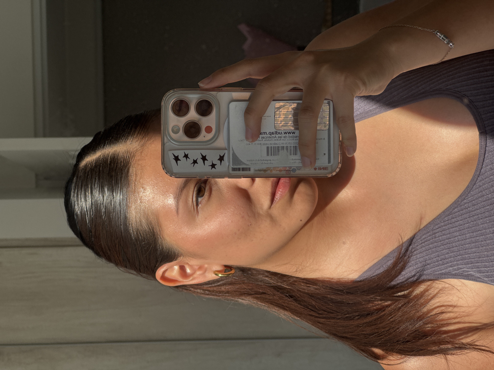 A close-up, vertically oriented selfie captures a person, likely female, holding an iPhone in a clear case. Her face is partially visible, showing her right eye, nose, and mouth, with her dark hair slicked back near the scalp. She has freckles on her cheek and wears a gold-toned hoop earring on her left ear. A delicate silver bracelet with cylindrical beads adorns her right wrist. She is dressed in a ribbed, possibly gray or purple, tank top.
The image is taken indoors during the daytime, characterized by strong, directional sunlight that creates prominent highlights on her skin and hair, and distinct shadows across her body and the background. The background itself is dark and indistinct.
Inside the clear phone case, which is adorned with small black star stickers, an identification card or similar document is visible. The text on this document appears reversed due to the reflection. Visible elements include "xm.qslbu.www", "Агвопета 261 eb bsbian", sequences of numbers such as "(553) 550 5000 | 1999 200 20210 800", and words like "141001 ob butisiloe", "boroug 65.10", "EX HICIE", and "IME". These details suggest an official document, possibly a driver's license or ID card from Mexico, given the provided context of San Francisco Ocotlán.
The activity depicted is the person taking a self-portrait using their phone's camera, likely by reflecting it off a mirror or similar surface.
