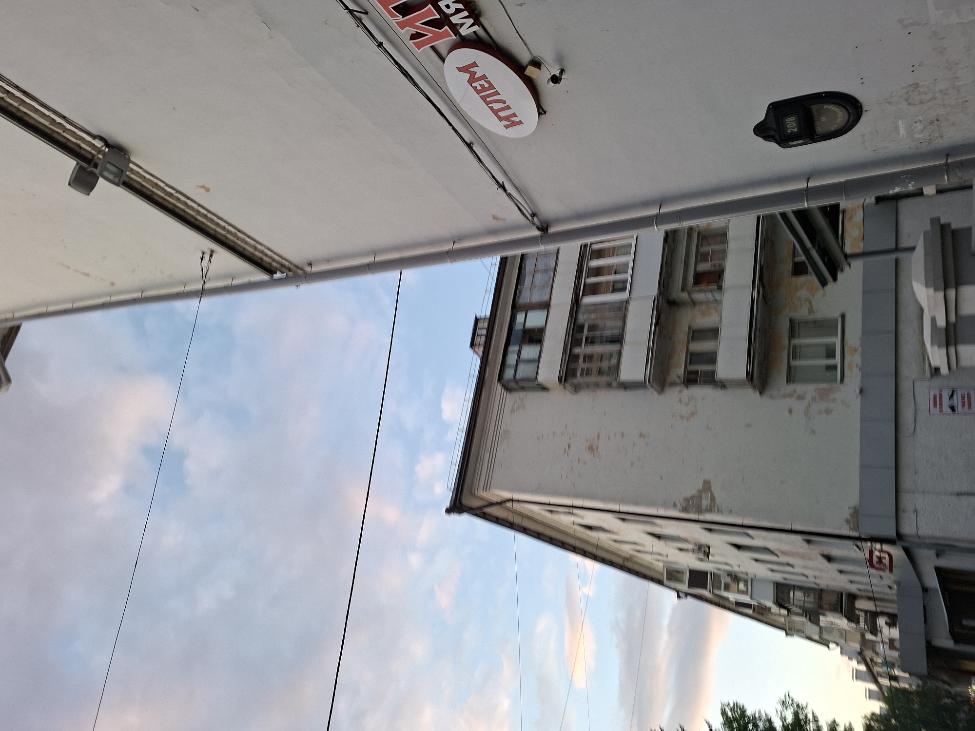 The image captures an outdoor urban scene, likely in Kazan, Russia, with a strong architectural presence and a backdrop of a partly cloudy sky. The perspective is from below, looking upwards, with the sky occupying a significant portion of the frame.
Dominating the upper part of the image is the underside of an awning or a similar structure, painted white. A metal bar runs along its length, with what appears to be a lighting fixture attached. A small, oval sign with red lettering in Russian is affixed to the underside of the awning. The text on the sign is partially obscured but reads "МИЛЛЕНИУМ" (Millennium).
Below the awning, an apartment building rises into view. The building's facade is a light, weathered color, with visible signs of age and some peeling paint. Rows of windows are visible, some with balconies. The building's architectural style suggests it is from the Soviet era.
The sky is a soft blue, interspersed with fluffy white and pinkish clouds, indicating it might be late afternoon or early evening. Two thin, dark wires run diagonally across the sky, suggesting the presence of overhead power lines or communication cables, common in urban environments.
A black, oval-shaped object, possibly a security camera or a sensor, is mounted on the underside of the awning, near the sign. Another similar object with a display showing "2016" is mounted further along.
The overall impression is a slice of everyday urban life, focusing on the interaction between built structures and the natural sky, with subtle hints of text and technology present in the scene. The low angle emphasizes the verticality of the buildings and the expanse of the sky.