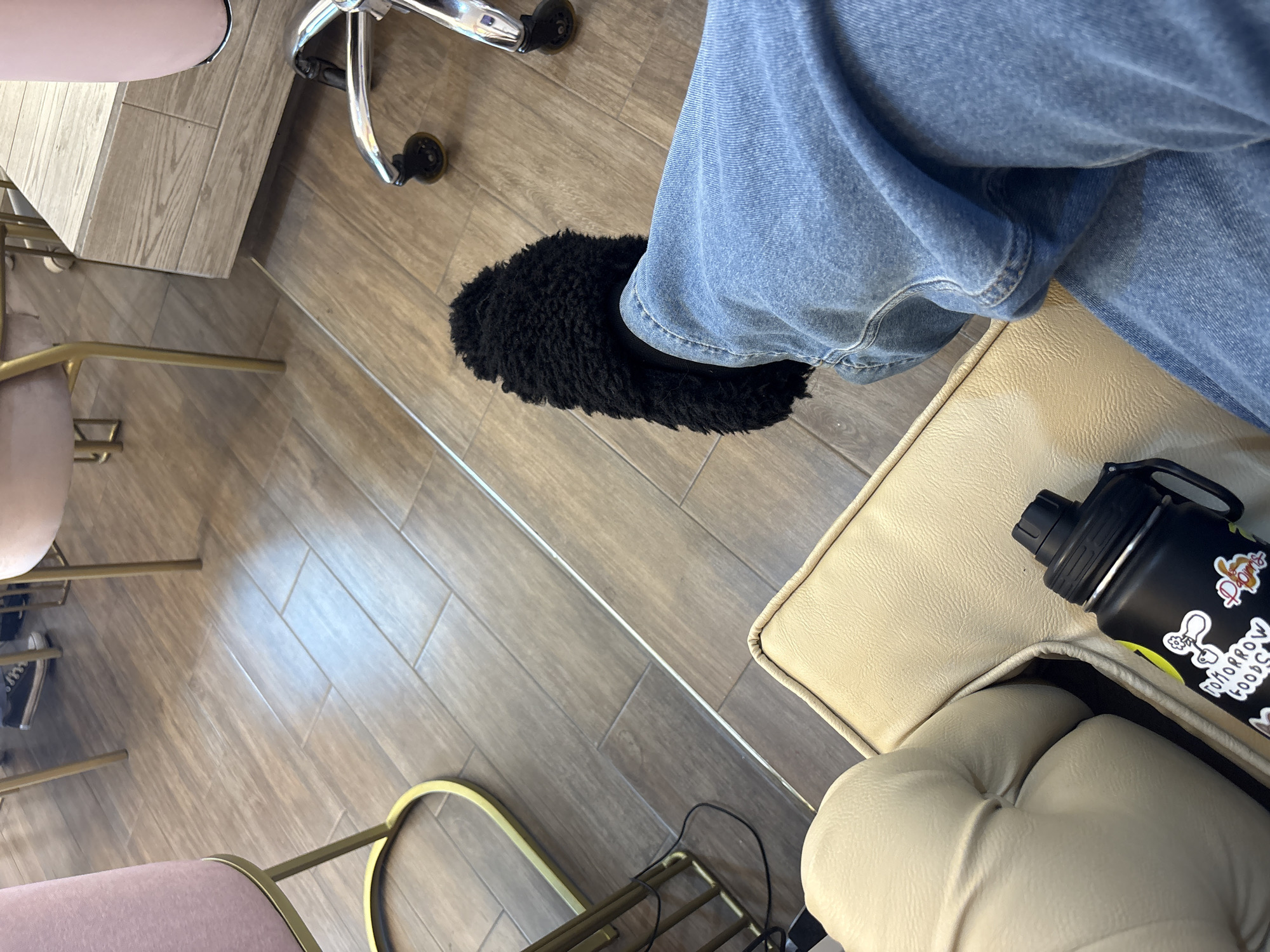A first-person perspective shot shows a person sitting in a chair with light blue jeans and a black fuzzy slipper on one foot. The person is seated on a light tan cushioned chair. A black water bottle with stickers is placed on the chair next to the person. The floor is made of light brown wood planks. A section of a desk or countertop is visible on the left side of the image. Part of an office chair with silver legs and black wheels is also visible in the upper left corner. A pink stool with gold legs is on the bottom left corner. The location is not identifiable from the image alone.