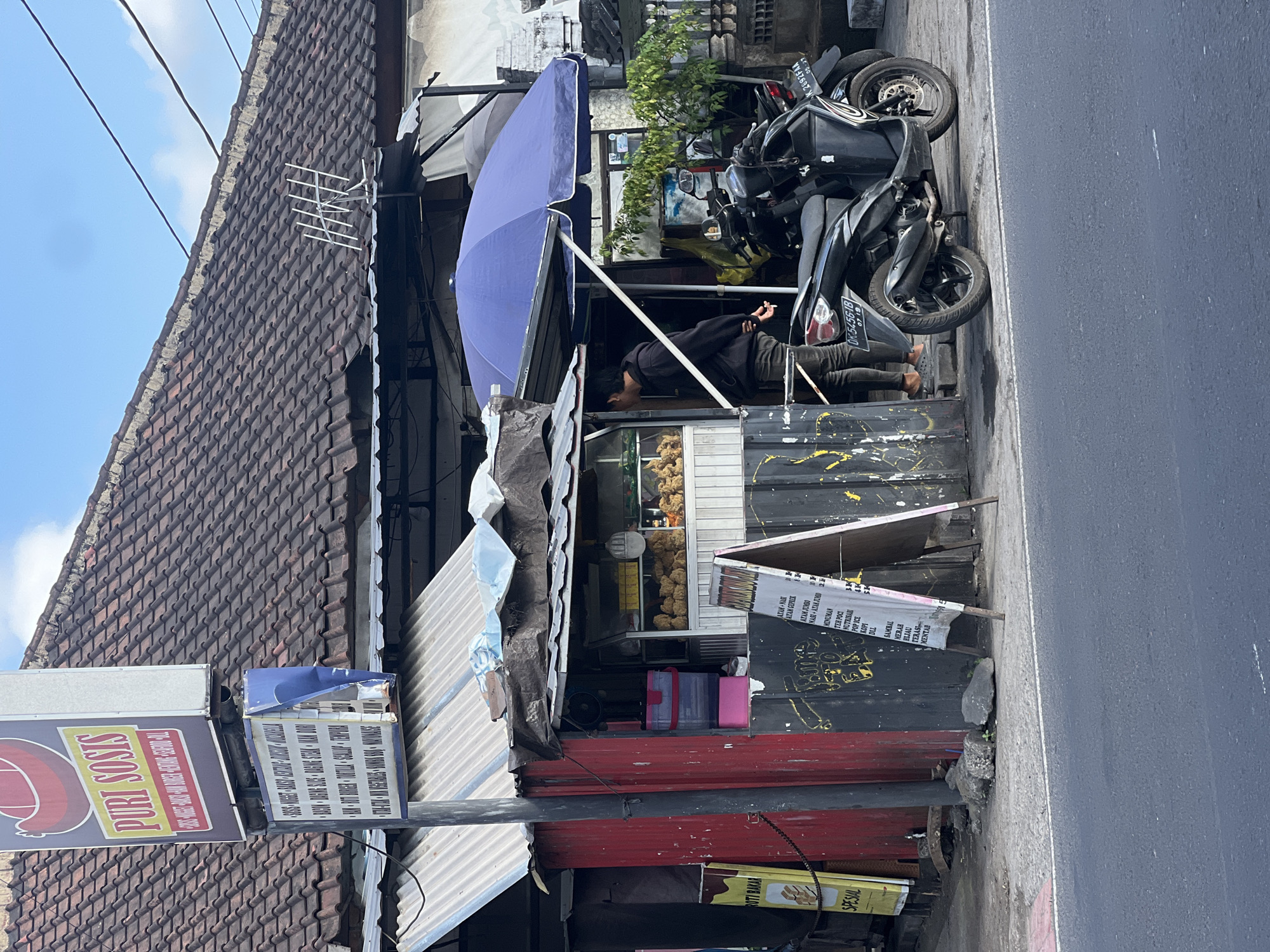 The image captures a street-side food stall in Denpasar, Indonesia, featuring a display of fried food items. A young man, wearing a black hoodie and dark pants, stands at the counter, likely a customer or employee. Two motorcycles are parked in front of the stall, suggesting a common mode of transportation in the area. The stall itself is modest, with a corrugated metal roof and a red-painted wall. A large blue umbrella provides shade over part of the seating area. The background shows a traditional tiled roof and some vegetation, indicating a residential or mixed-use urban environment. A sign above the stall advertises "Puri Sosis," along with a list of food and beverage options. The overall scene depicts a typical everyday commercial activity in an Indonesian city.