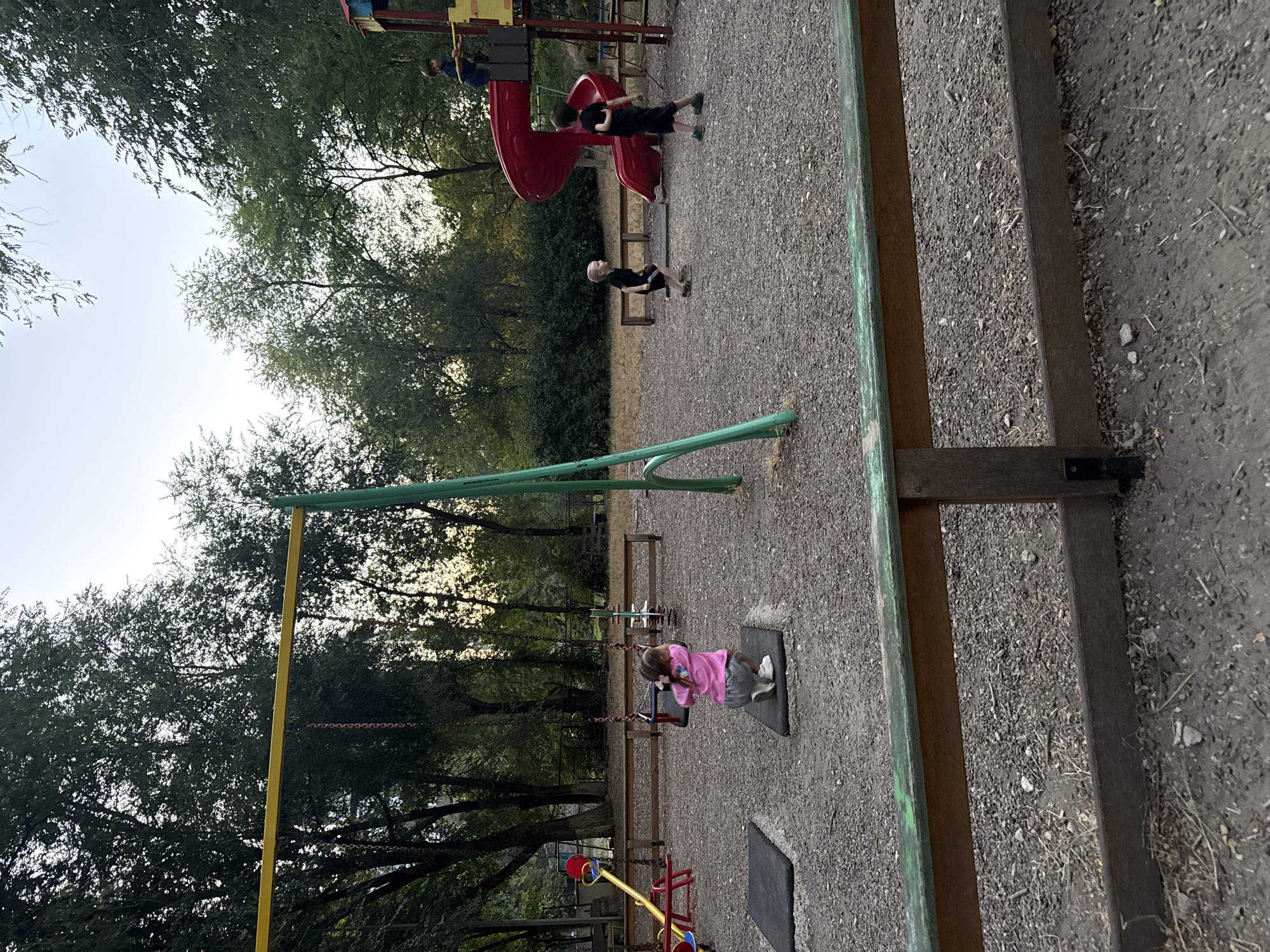 This image depicts an outdoor playground scene. In the foreground, a wooden fence with a weathered green top rail is visible, separating the viewer from the playground. The playground surface is covered in small gray gravel and wood chips.
Dominating the center of the image is a swing set with a yellow horizontal bar supported by green poles. One swing seat is occupied by a young child wearing a pink shirt and grey shorts, who is in mid-swing. To the left of the swing set, a red and yellow seesaw-like structure is partially visible.
In the mid-ground, to the right, is a red plastic slide that is part of a larger playground structure. A young child in a blue shirt is standing at the top of the slide, holding onto a yellow bar. Another child in a black shirt and shorts is running towards the slide. Further to the right, a child in a black shirt and shorts is standing near the slide. Several wooden benches are placed throughout the playground area.
The background is filled with dense green trees, suggesting a park or wooded area. The sky is visible through the trees, appearing overcast or at dusk. The overall impression is that of a public park playground. No specific landmarks or identifying features for Chișinău, Moldova, are evident within the image itself.