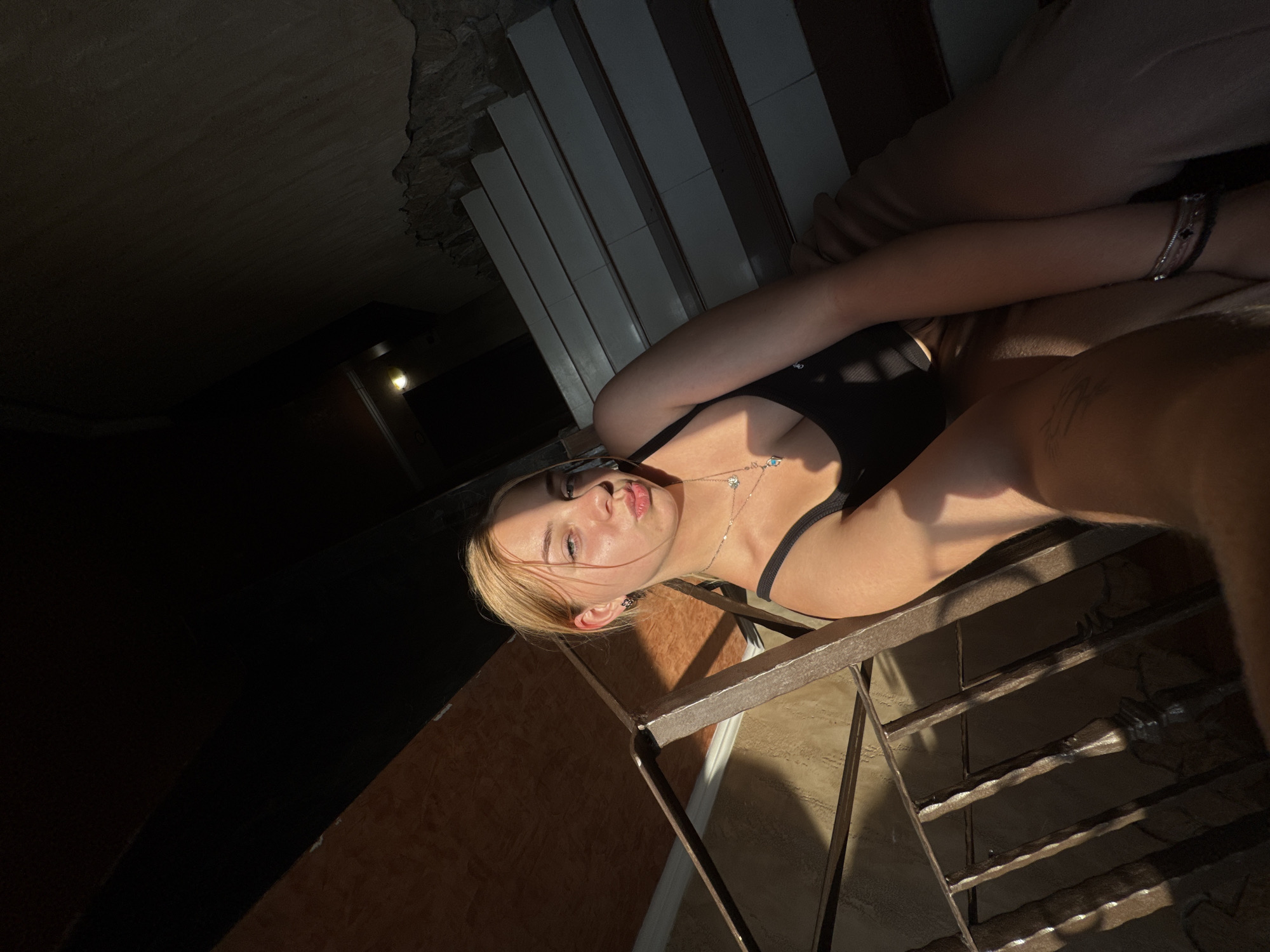 In the city of Sredets, Bulgaria, a young woman with blonde hair pulled back is taking a selfie on a staircase. She is wearing a black tank top and tan pants, and a silver necklace with a small pendant. The lighting suggests it is late afternoon, with sunlight casting shadows across the scene. The staircase appears to be made of concrete with white risers and darker treads, bordered by a metal railing and textured walls. The background shows an interior space, possibly a residential building or a public area.