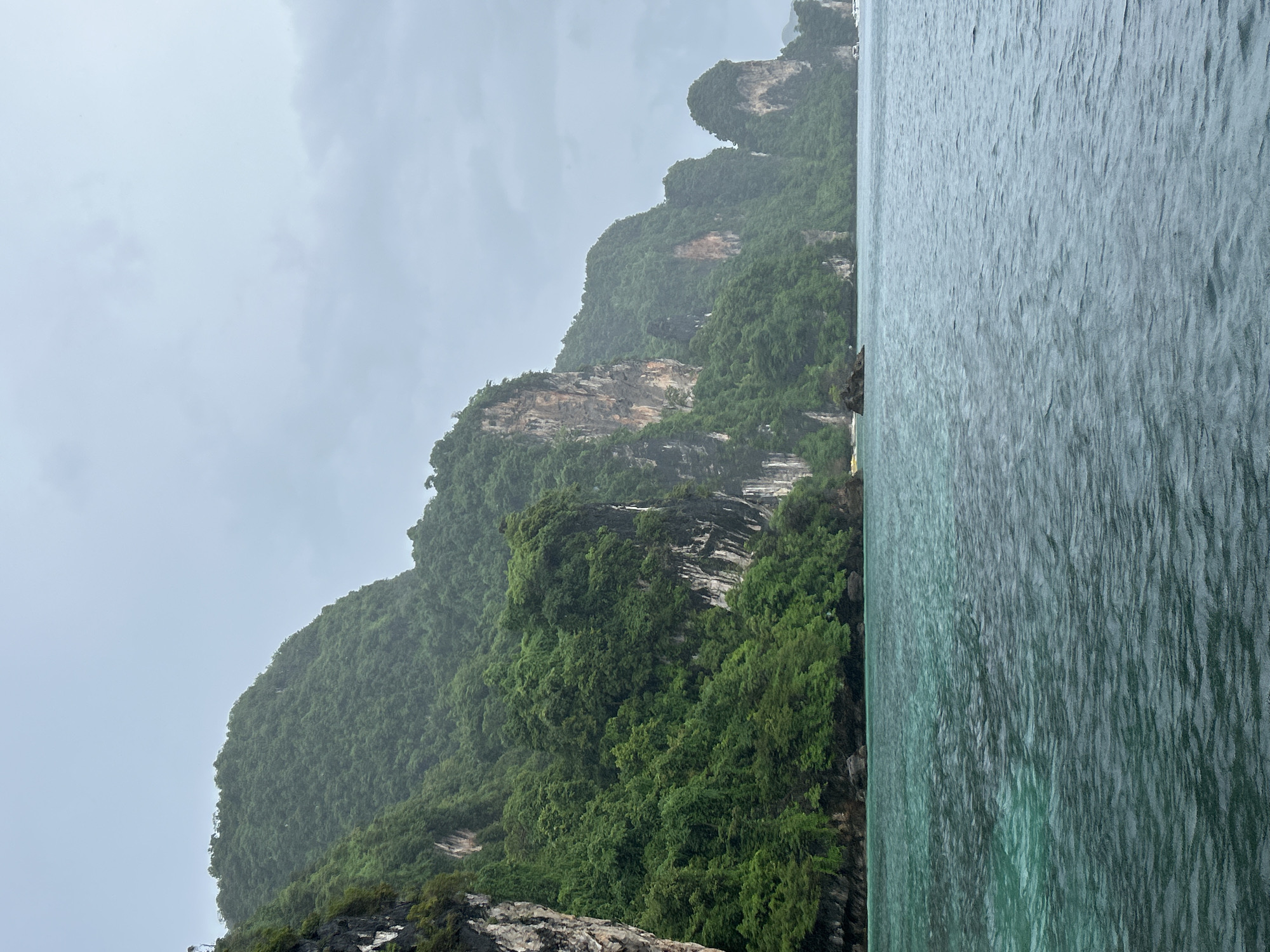 The image depicts a scenic view of limestone karsts covered in lush green vegetation, rising dramatically from the turquoise waters of the sea. The karsts are characterized by their steep, rugged cliffs and dense foliage. In the foreground, the water exhibits a gradient of colors, shifting from a clear, aquamarine hue near the shore to a deeper, darker blue further out. The sky is overcast with a pale, grayish hue, suggesting a cloudy or potentially rainy day. The scene conveys a sense of natural beauty and tranquility. The specific location is identified as Haad Hin Khom, Thailand.