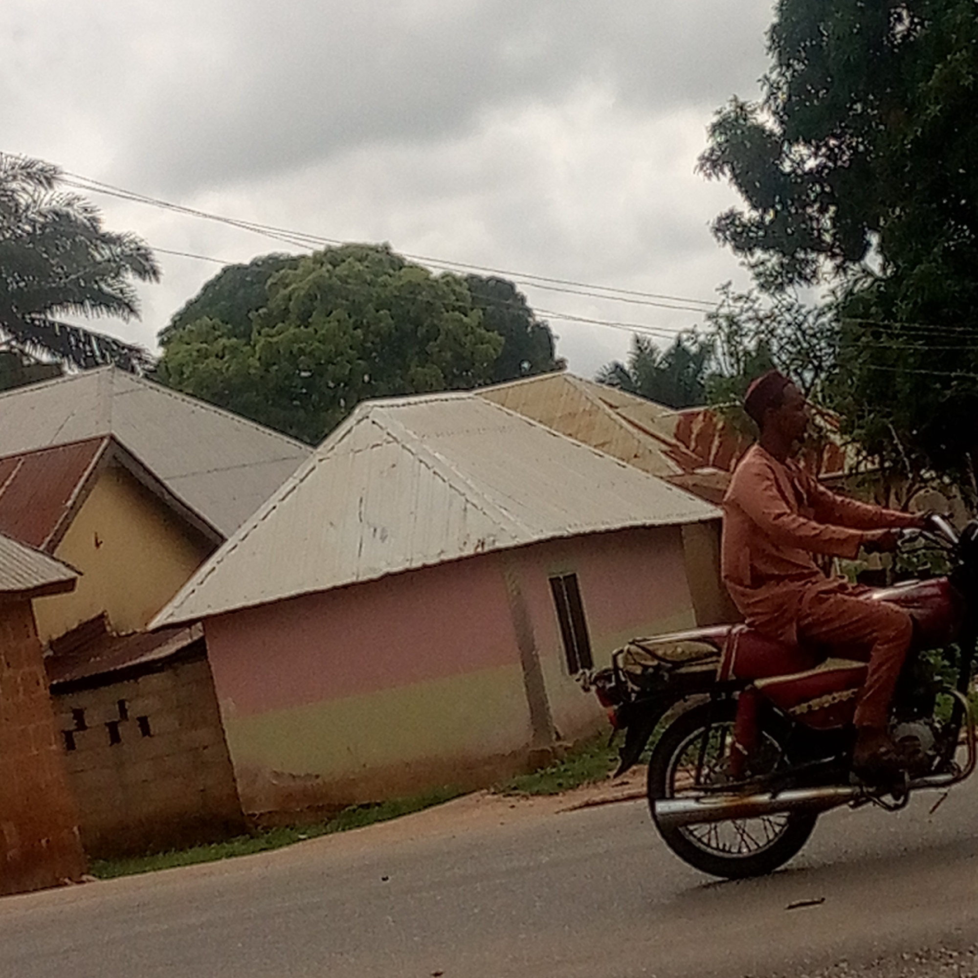 The image depicts a man riding a motorcycle on a paved road in Mararaba, Nigeria. The man is wearing a reddish-brown outfit and a maroon fez hat. The motorcycle is also reddish-brown. Behind the motorcycle are several houses with corrugated metal roofs. The house closest to the motorcycle has pink and tan painted walls. The sky is overcast. Vegetation, including large trees, is visible in the background. Powerlines are visible overhead.