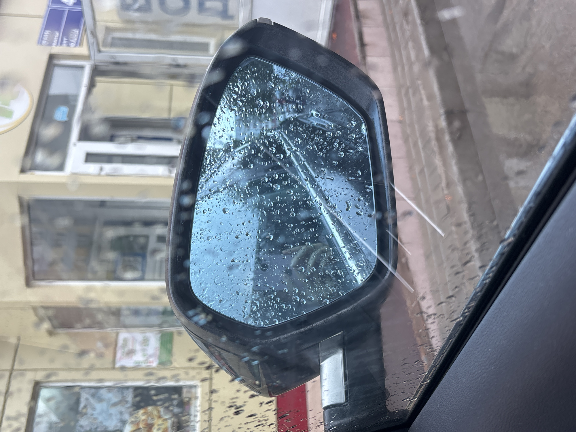 The image shows a car's side mirror with raindrops on its surface. The mirror reflects a street scene, possibly a commercial area with buildings and doorways. The foreground shows the interior of the car, including part of the dashboard and window. The reflection in the mirror is somewhat distorted by the raindrops, but it appears to be daytime, and the weather is rainy. The overall impression is that of a rainy day in a city. There is no specific indication of the city of Naberezhnye Chelny, Russia.