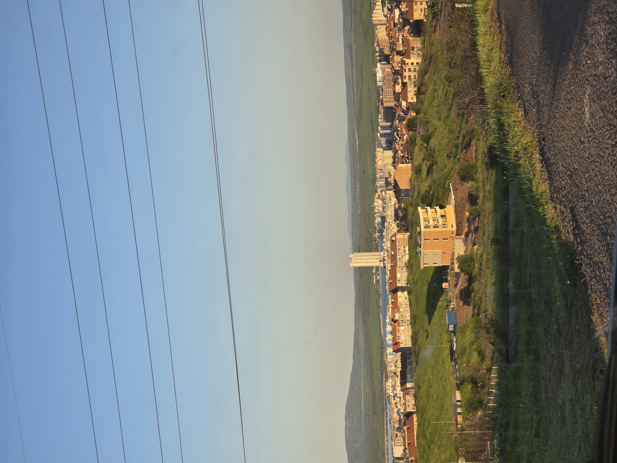 The image presents a panoramic landscape view of a city, likely Burgas, Bulgaria, captured from an elevated perspective. The upper portion of the frame features a clear, pale blue sky, horizontally traversed by several parallel utility wires that run diagonally upwards from left to right.
Below the sky, an expansive urban settlement stretches across the mid-ground and foreground. The city comprises numerous buildings, predominantly residential or commercial structures, displaying a mix of architectural styles and colors, with warm tones of beige, brown, and white being common. A particularly tall, slender white building stands out prominently in the middle distance, surrounded by other structures.
The urban area is interspersed with and borders natural elements. Lush green hills and slopes, covered with grass and scattered trees, form a significant part of the landscape, providing a verdant contrast to the built environment. A narrow body of water, possibly a canal or a part of a larger aquatic feature, is visible running horizontally through the scene, separating portions of the town from the green terrain.
On the far right side of the image, a dark asphalt road or path is partially visible, suggesting the viewpoint is from alongside such a surface. The lighting in the scene, characterized by a soft, warm glow on the buildings and elongated shadows cast by objects and terrain, indicates either early morning or late afternoon. The sky is clear, suggesting fair weather. There is no discernible human activity or visible text within the frame, contributing to a serene and observational atmosphere.