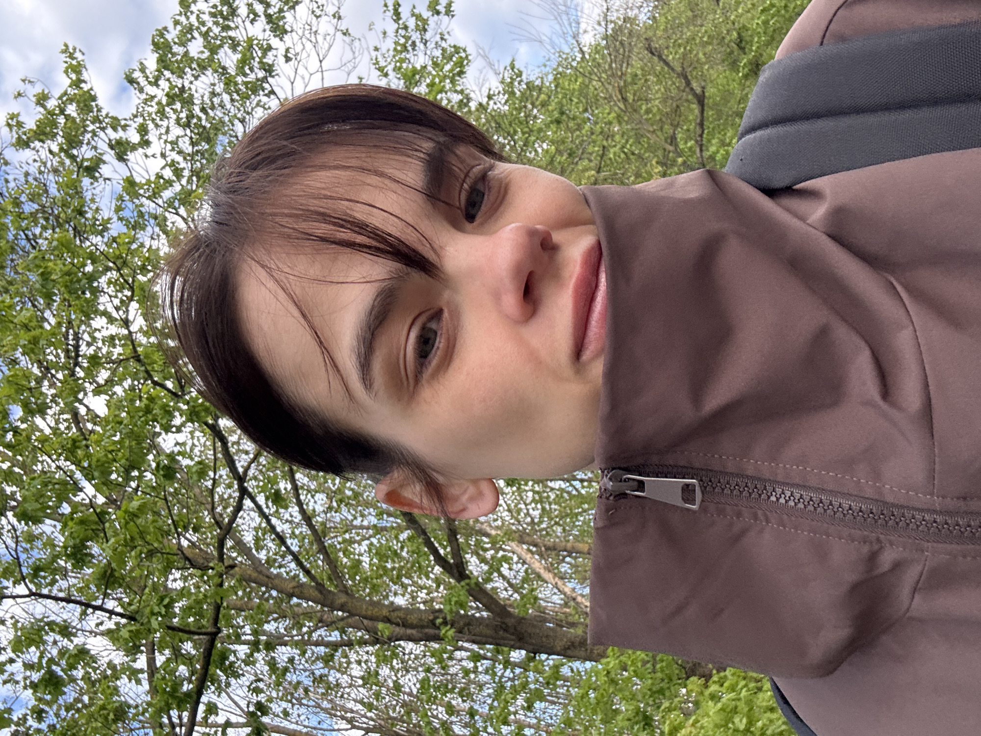 A close-up, slightly tilted portrait captures a young individual, likely female, from the chest up, looking directly at the camera. The person has short, dark brown hair with bangs lightly brushed across their forehead, fair skin, and dark eyes. Their expression is neutral and calm. They are dressed in a brown jacket, visible on the right side of the frame, featuring a metal zipper pull and teeth. A black, padded strap, consistent with a backpack, is visible over their right shoulder.
The background is an outdoor scene dominated by numerous deciduous trees with fresh, vibrant green leaves, indicating spring or early summer. Some thinner branches without dense foliage are also visible against a bright, cloudy sky. The lighting is soft and diffused, typical of an overcast day. The setting suggests a park or natural environment, aligning with locations found in Rostov-on-Don, Russia. There is no visible text in the image.