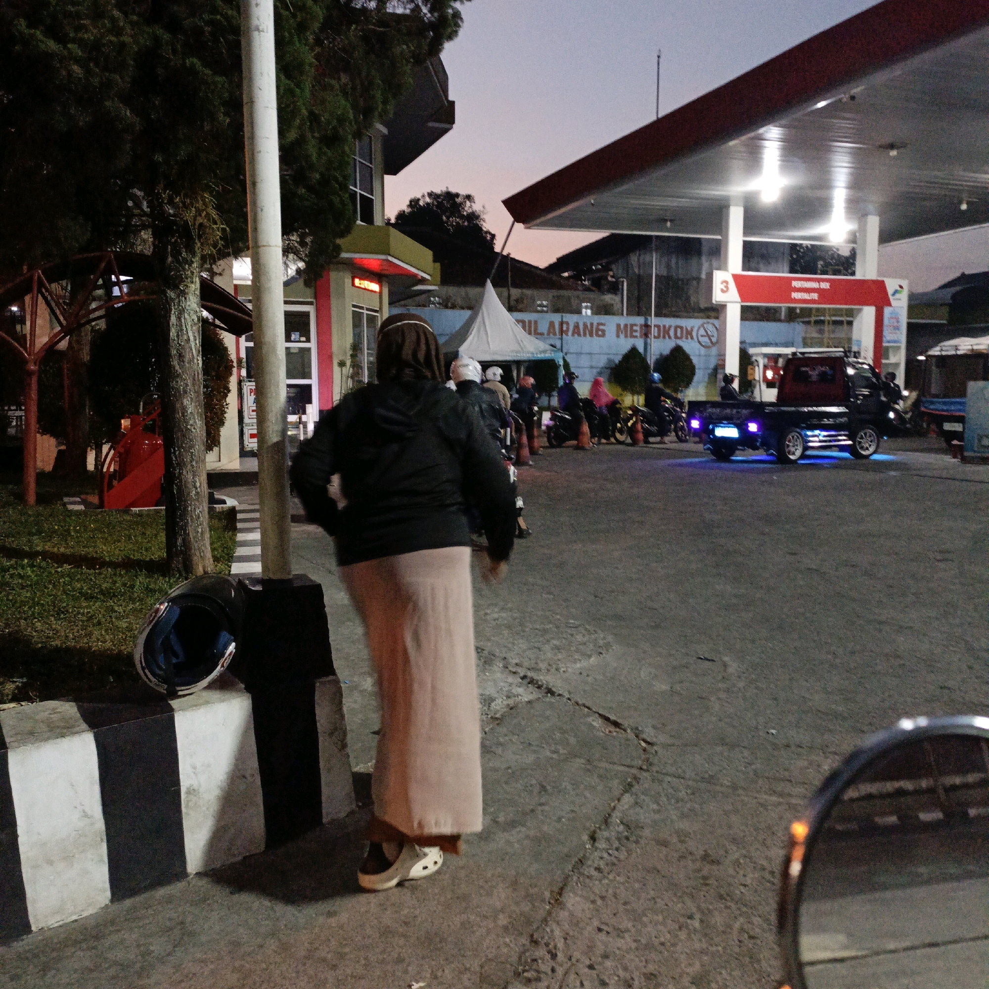 The image depicts an outdoor scene at a gas station in Salatiga, Indonesia, at dusk. In the foreground, a person wearing a brown head covering, a black jacket, and a long pink skirt is walking away from the camera. A black motorcycle helmet rests on a striped concrete barrier to the left. Behind the person, several motorcycles are parked, some with riders. A black pickup truck with blue underglow lighting is also present. The gas station has a red and white awning with lights, and a sign that reads "DILARANG MEROKOK" (No Smoking). The sky is a soft gradient of orange and grey, indicating twilight. The ground is paved with asphalt and concrete.