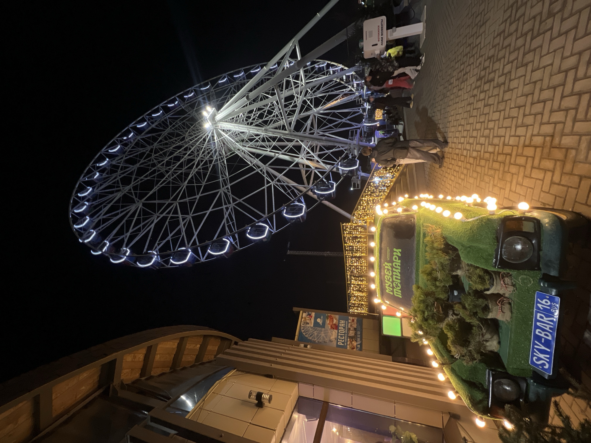 At night in Kazan, Russia, a Ferris wheel with white lights illuminates the sky. People stand on a brick pathway. A decorated vehicle with a "SKY-BAR 16 RUS" license plate and Christmas lights adorns it. A building with a sign reading "МУЗЕЙ ТОПИАРИ" (Museum of Topiary) is visible. Another sign advertises a restaurant.