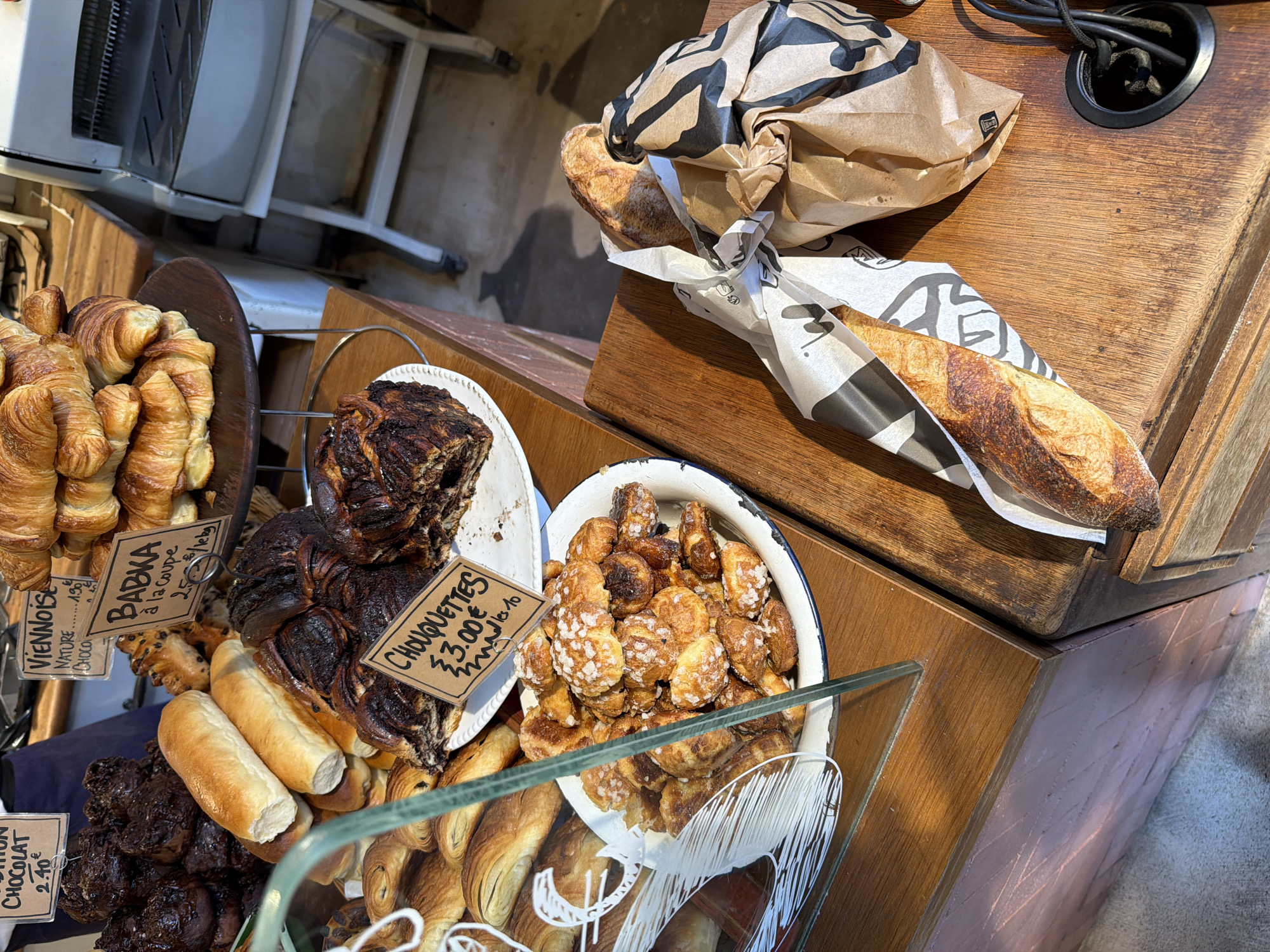 This is a shot of a bakery display case in Paris, France. In the display are a variety of baked goods, including croissants, babka, chouquettes, and pain au chocolat. The babka has a price tag of 2.50€/slice, and the chouquettes are priced at 3.00€/10. There is also a paper bag on top of a wooden counter, containing a baguette, or two baguettes. The paper bag is printed with a zebra stripe design. A power cord is also visible in the top right corner of the image. The scene is lit by natural sunlight.
