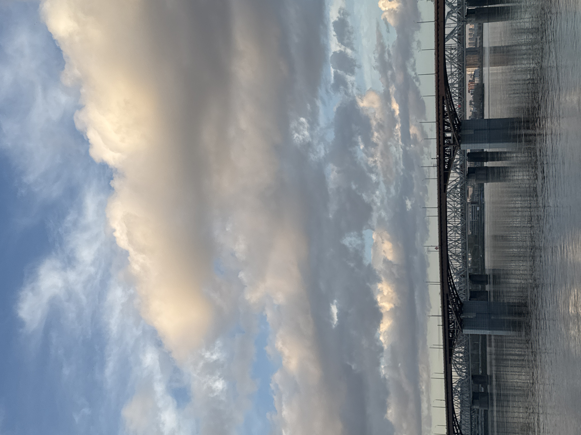 The image shows a bridge spanning a body of water. The bridge is constructed of steel and has a complex truss structure. Pillars support the bridge in the water. The water reflects the bridge and the sky. The sky is filled with clouds, with some areas of blue visible. The city of Krasnoyarsk, Russia, is not clearly visible.