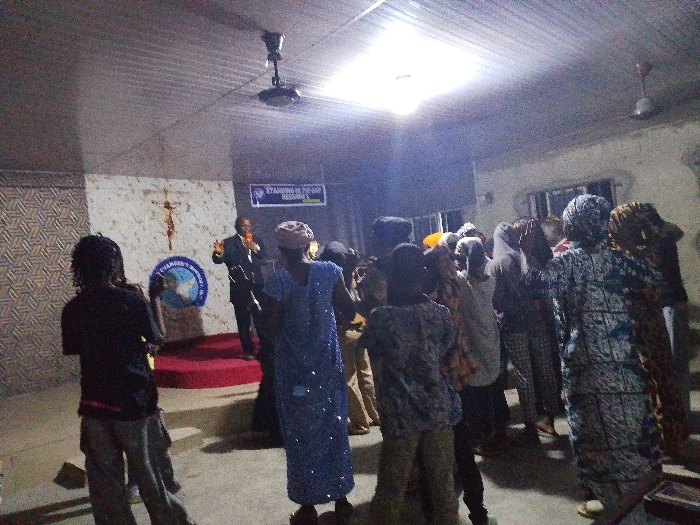 Inside a small church, a man in a suit stands on a red-carpeted stage, speaking into a microphone. Behind him is a wall with a crucifix and a church logo. In front, a diverse group of people, including women in patterned dresses and head coverings, face the speaker, seemingly in prayer or worship. Ceiling fans hang overhead, and a window is visible on the right wall. The room is illuminated by a bright overhead light.